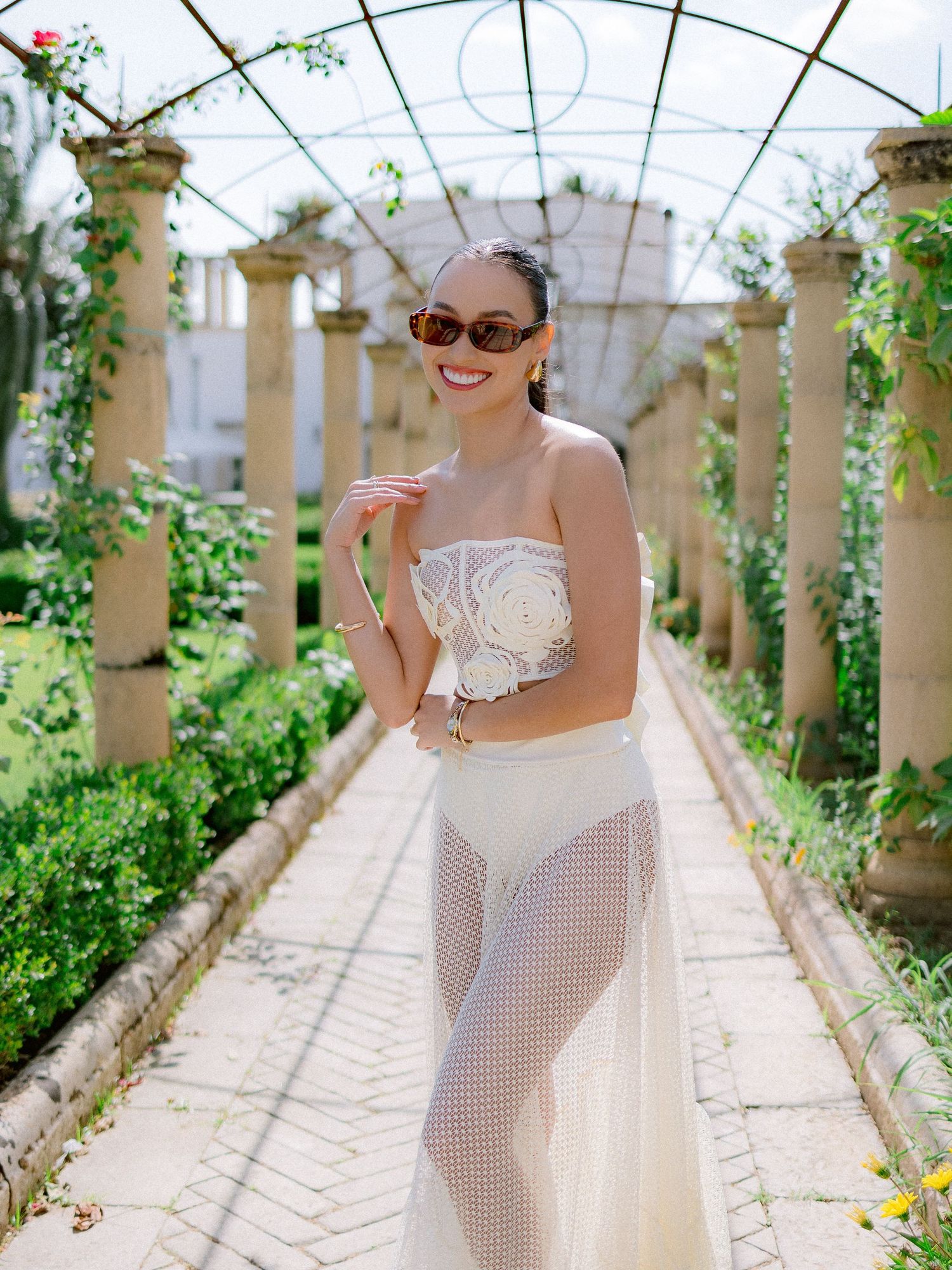 A strapless white lace dress flows elegantly in an outdoor garden pergola with stone columns and climbing vines.