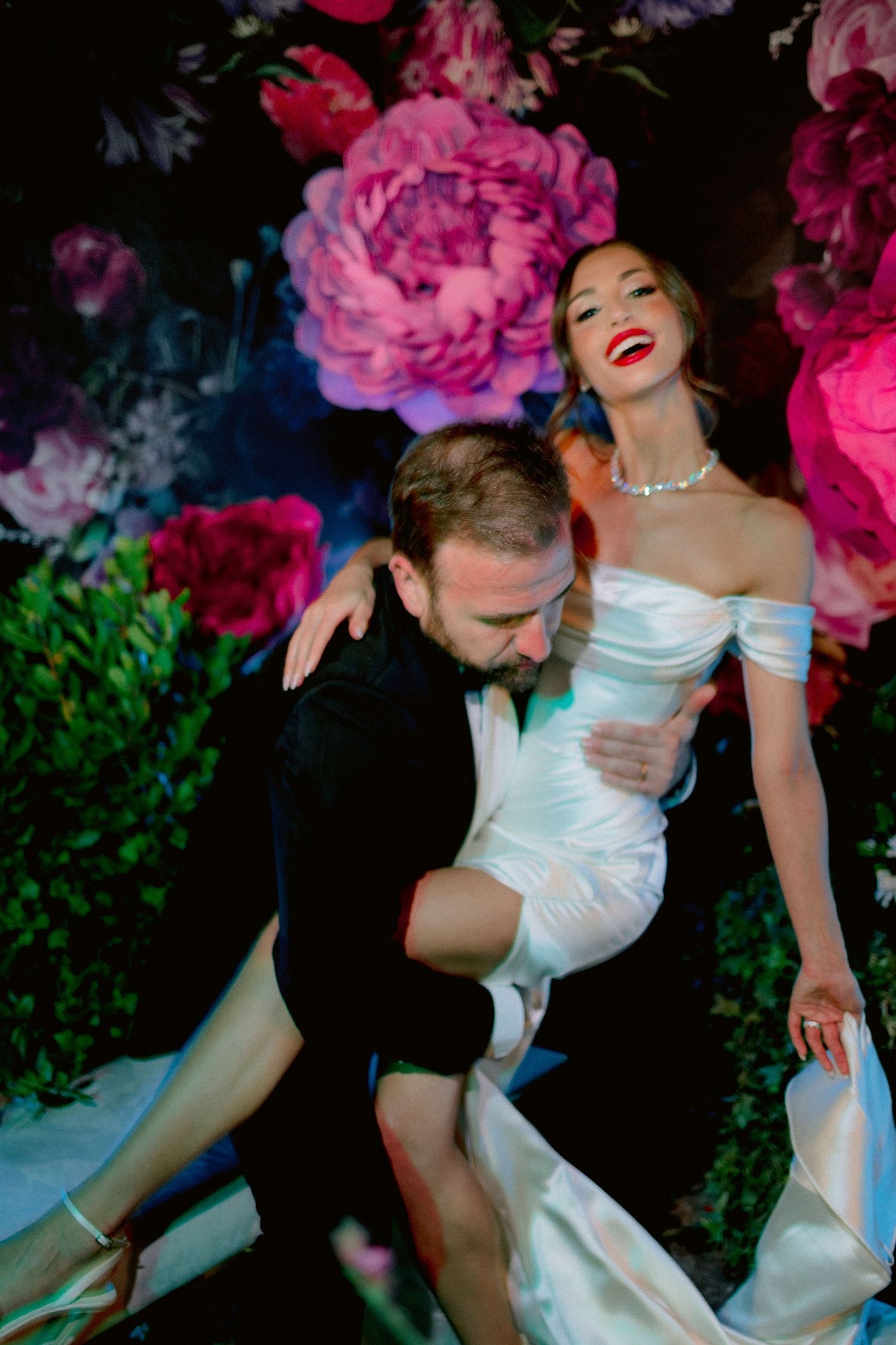A romantic couple poses in front of a floral backdrop with pink peonies, the bride wearing an elegant white off-shoulder gown.