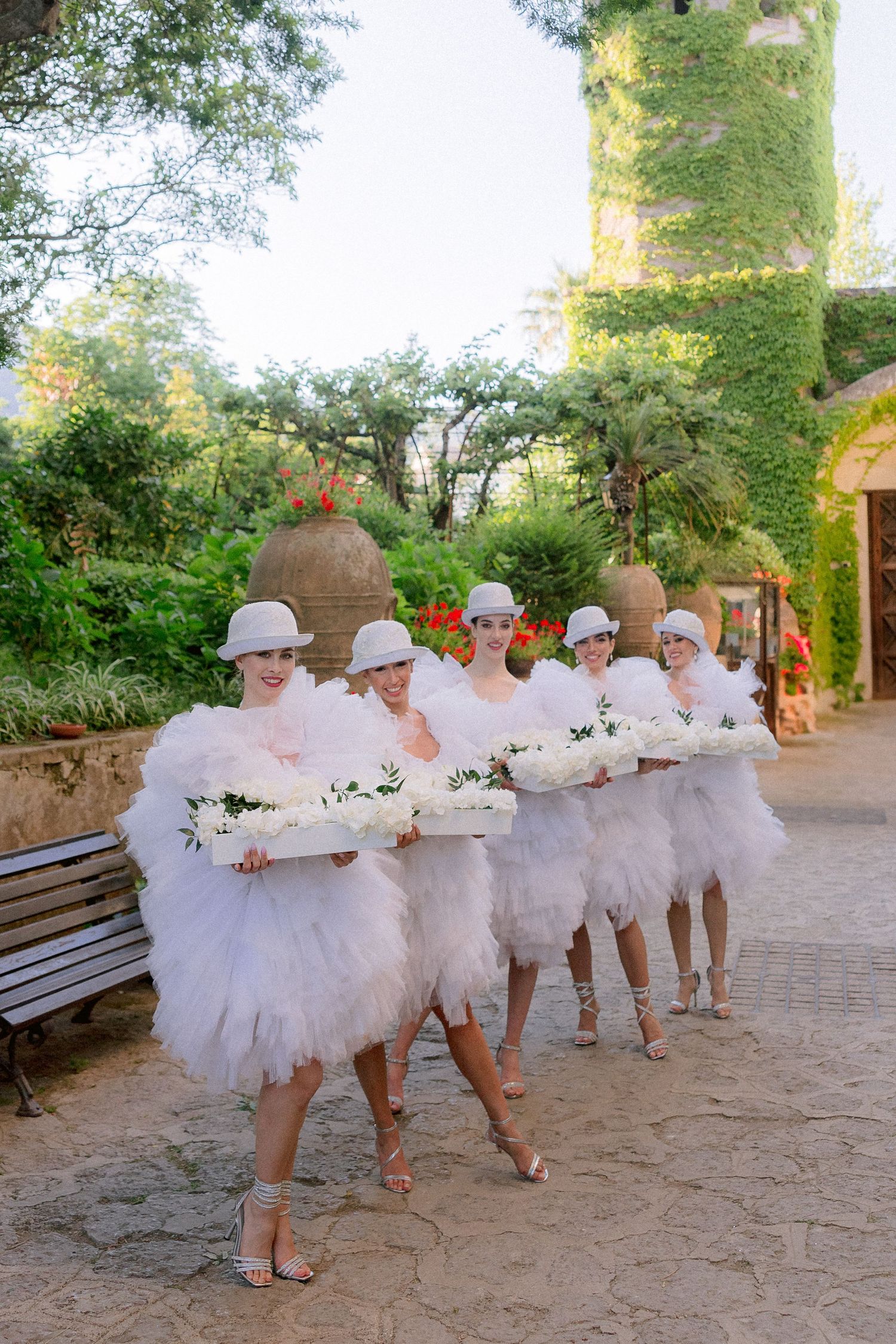 Dancers in white tulle tutus and matching hats carry floral arrangements while posing in a line on a garden path.