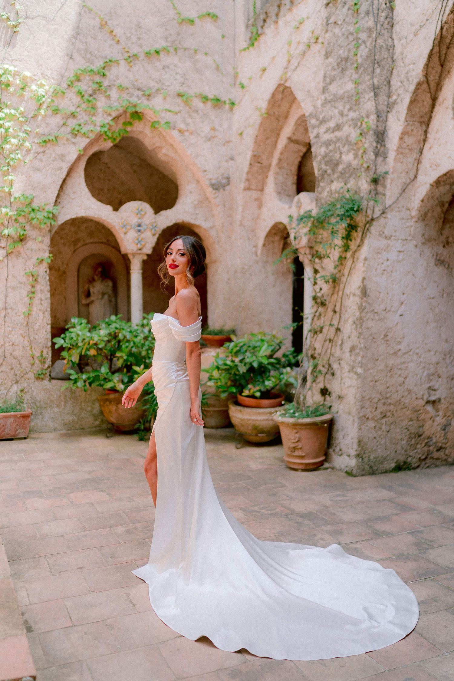 Elegant white wedding dress with long train photographed in an ancient Mediterranean stone courtyard with arched walkways.