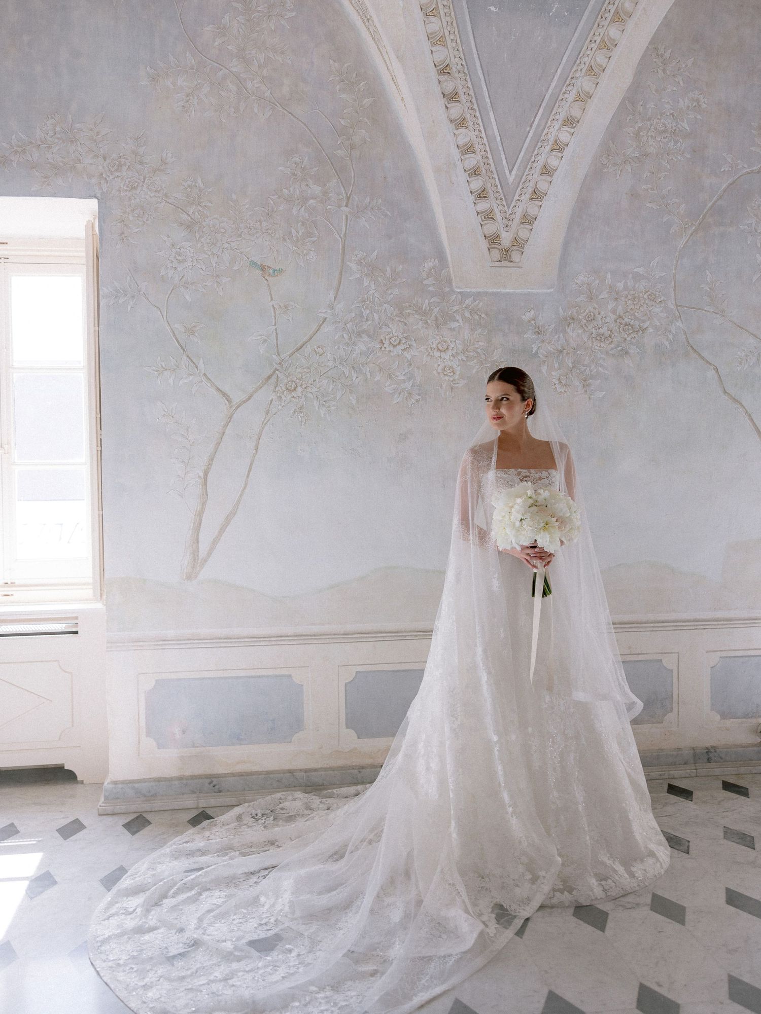 An elegant wedding gown with a flowing cape and long train photographed in a light-filled room with ornate wall details.