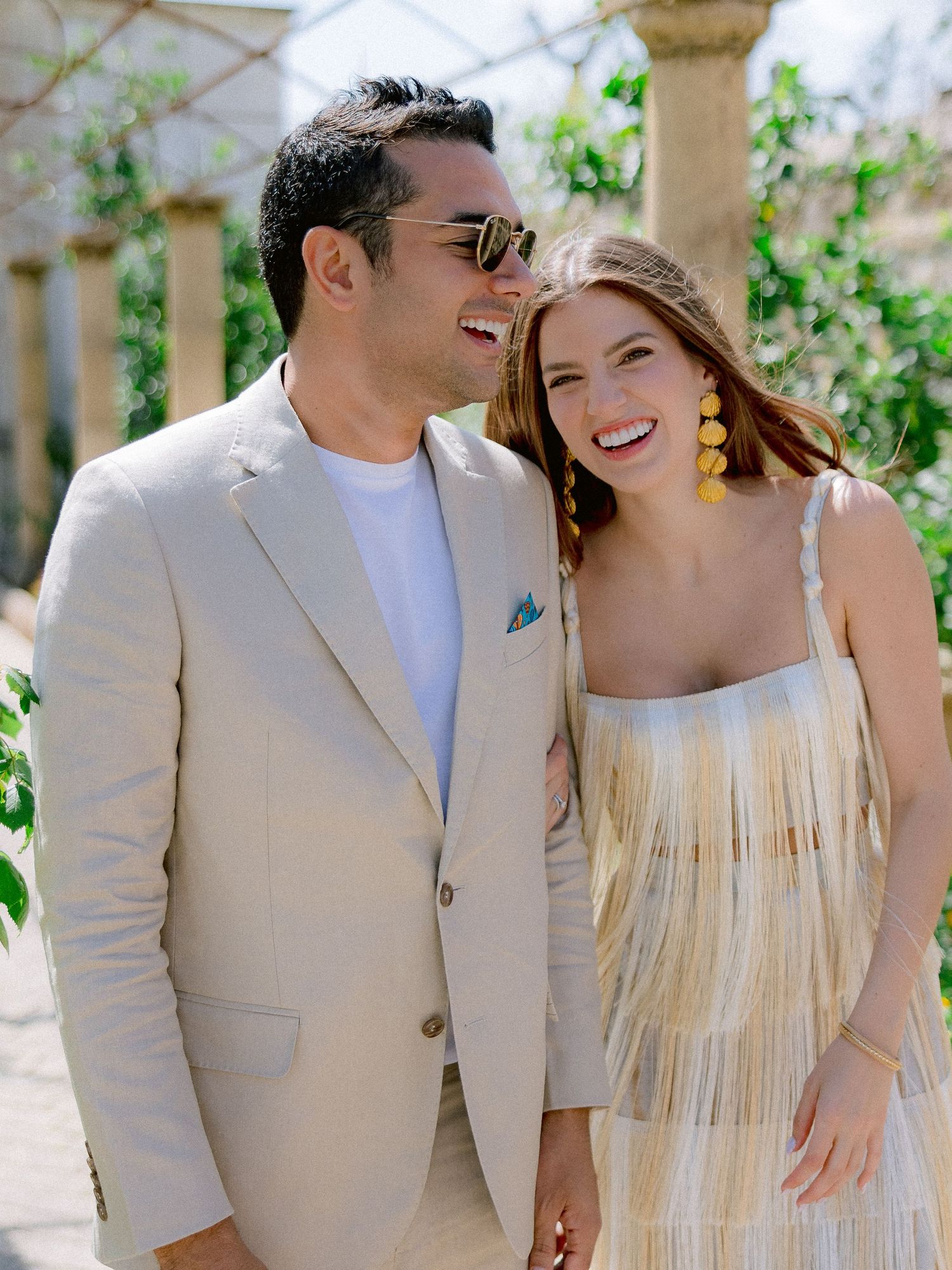 A couple smiles together outdoors, with the woman in a fringed dress and the man wearing a beige suit and sunglasses.
