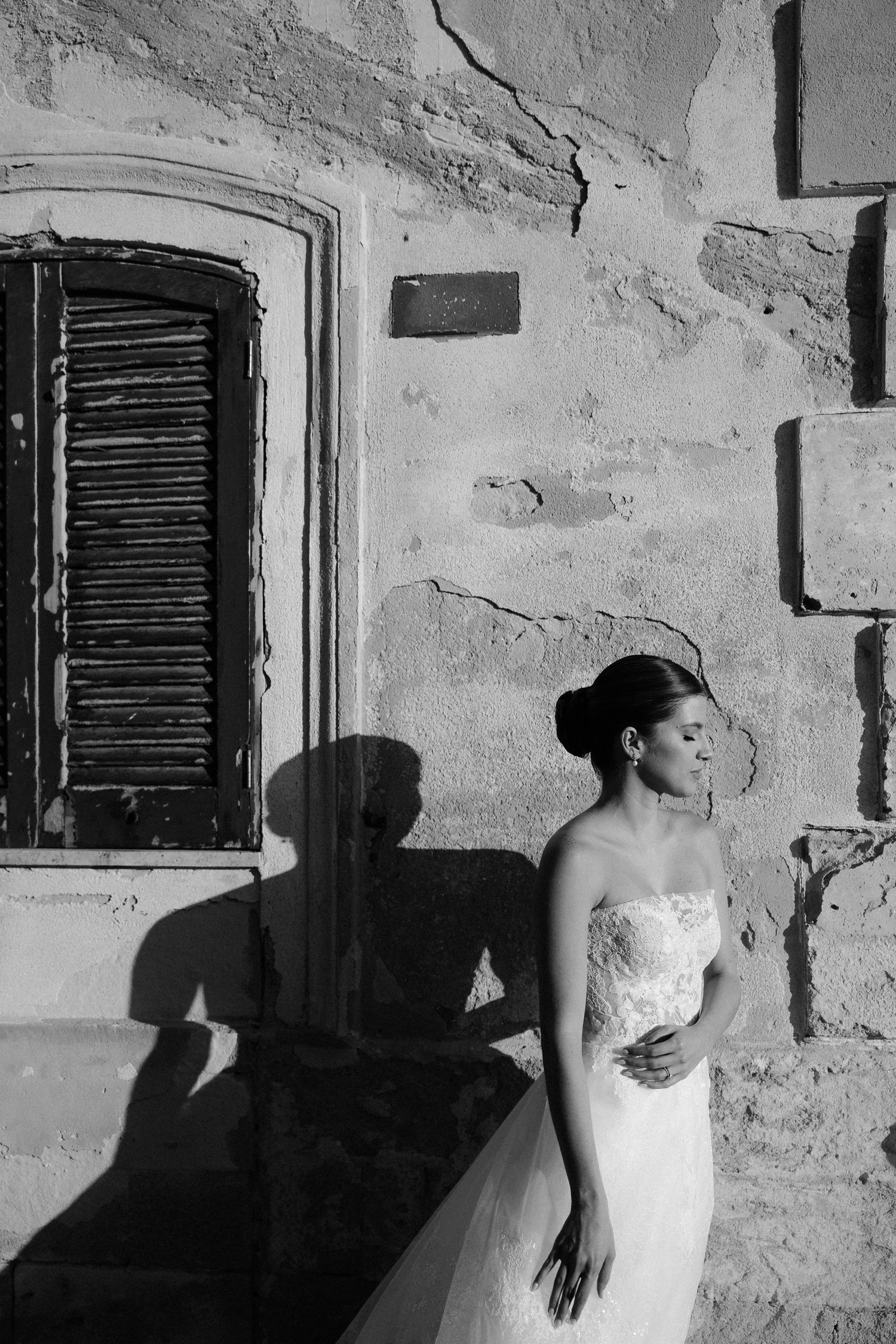 Black and white artistic wedding photo with dramatic shadow cast on textured stone wall beside shuttered window in salento, Puglia.