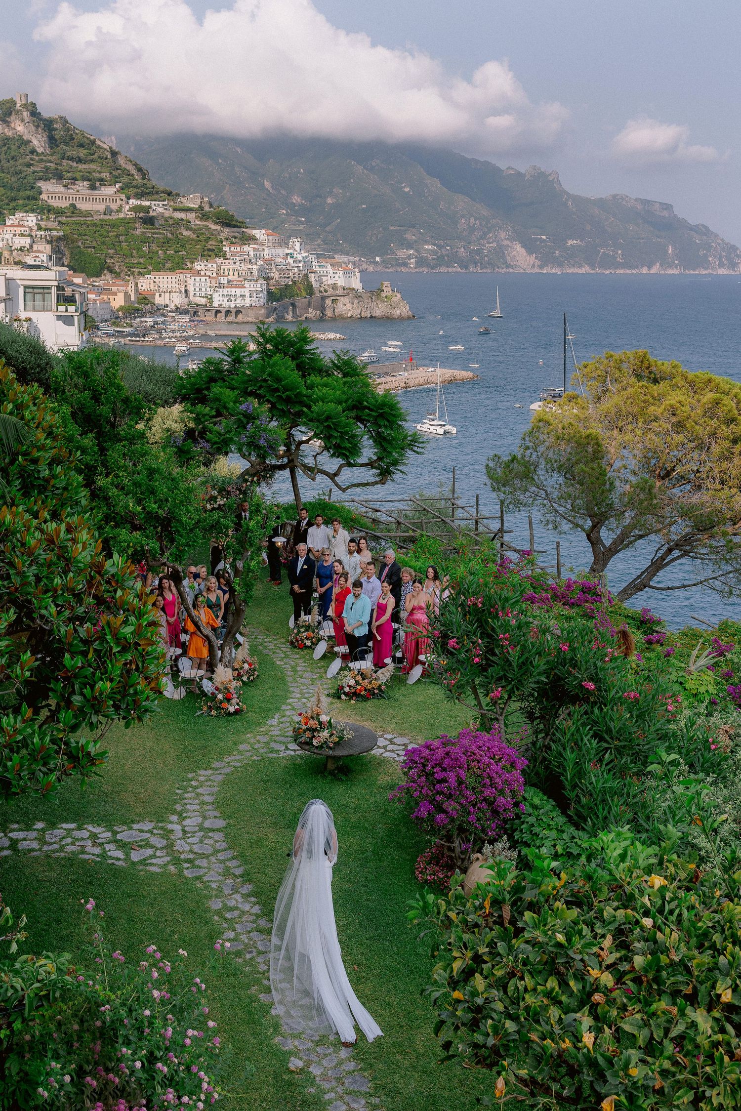 A wedding ceremony taking place in a lush garden overlooking the stunning Hotel Santa Caterina in the Amalfi Coast with boats in the sea.