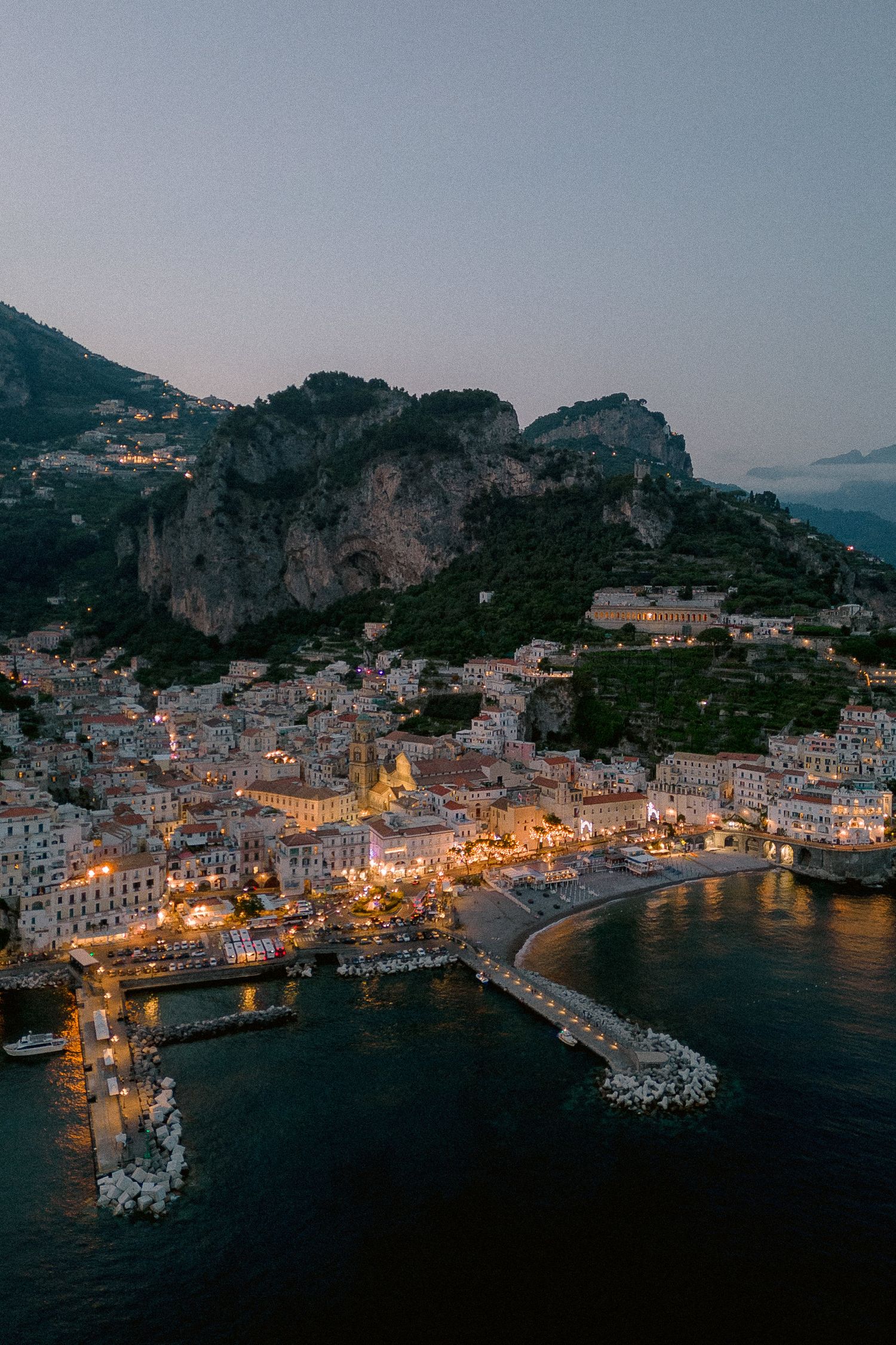 Aerial twilight view of Amalfi Coast in Italy showing illuminated coastal town nestled between mountains and Mediterranean Sea.