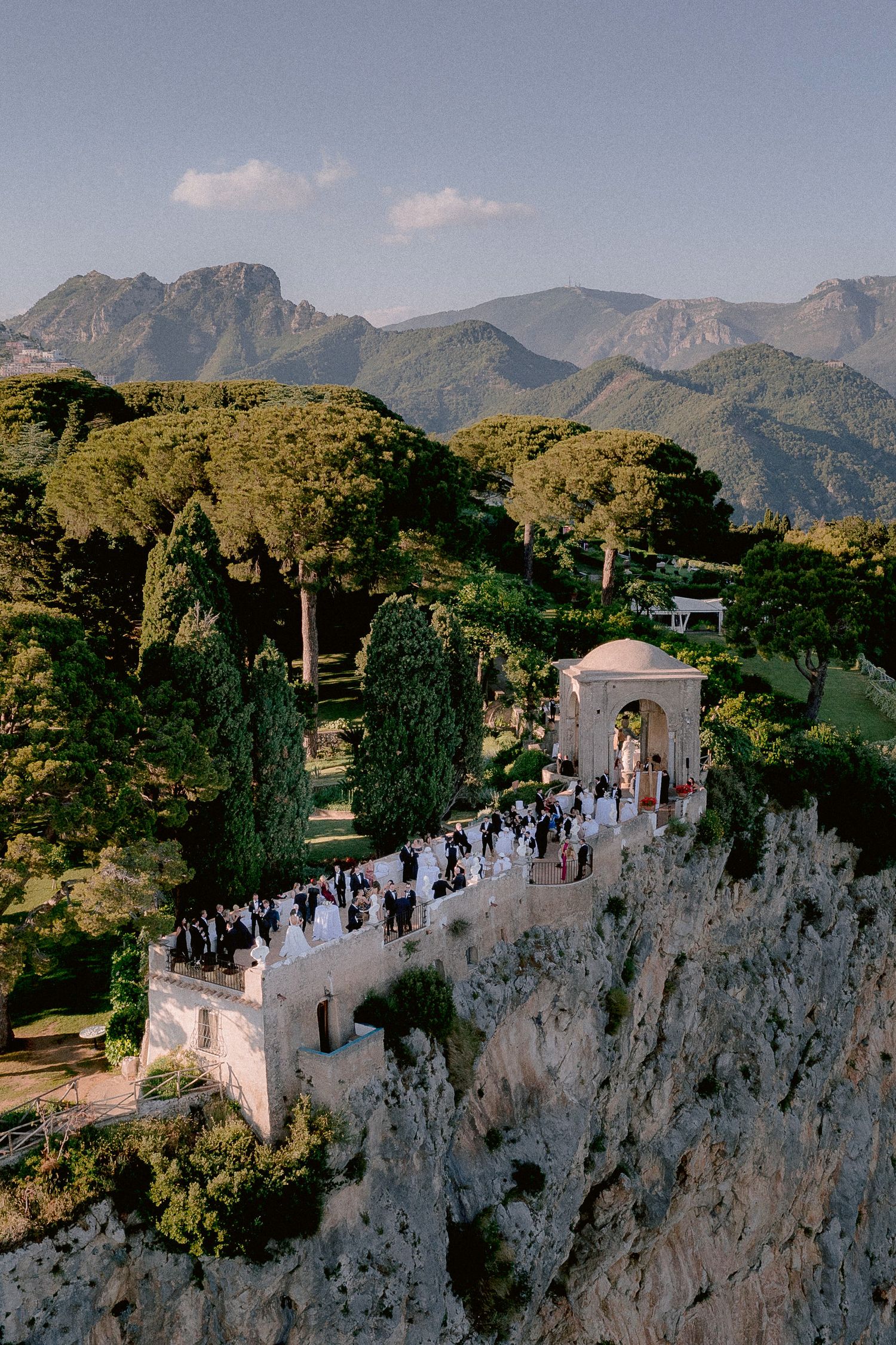Aerial view of a cocktail hour on a white cliffside terrazza dell'infinito at Villa Cimbrone overlooking mountains and trees.