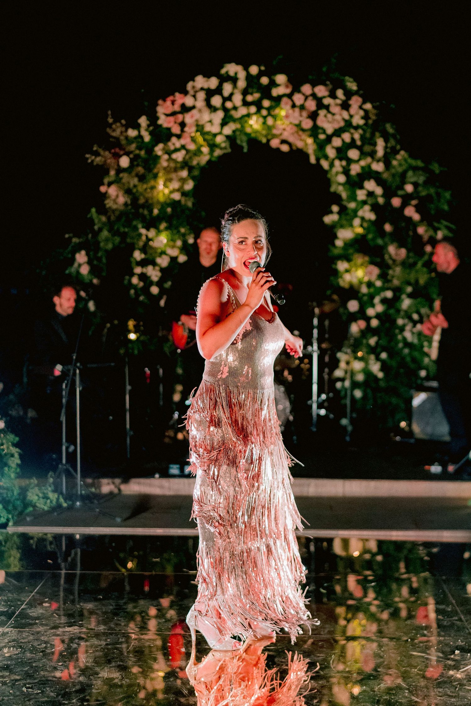 A performer in a flowing white fringed evening gown sings under a floral arch at an elegant nighttime event.