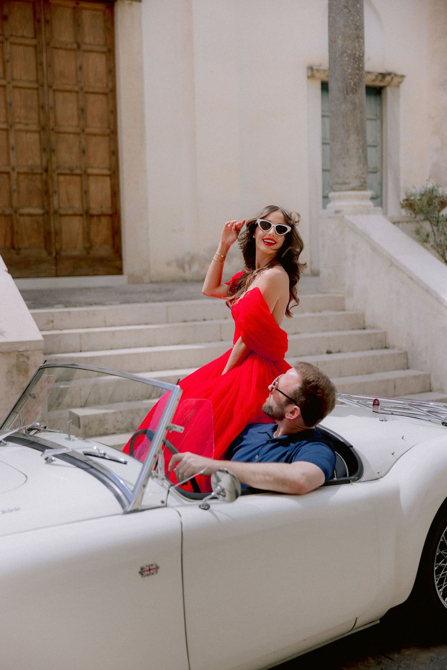 A couple in a vintage white convertible car, with a woman in a red dress posing by stone steps.