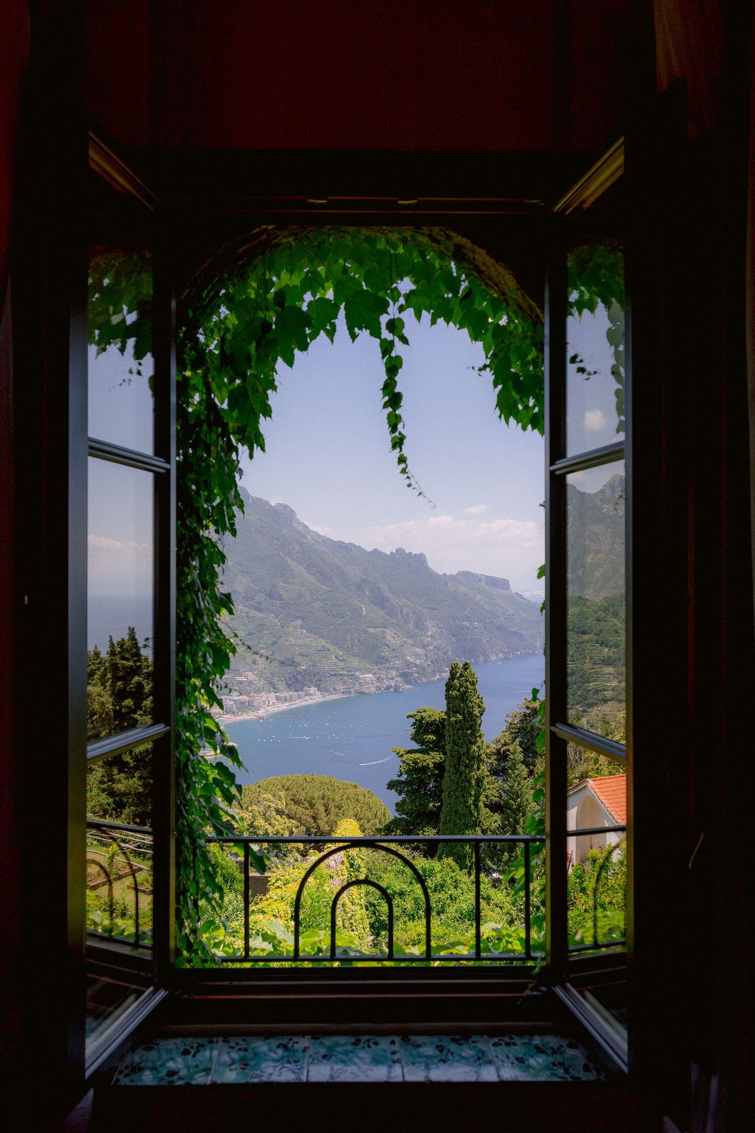 Open window with ornate iron railing framing scenic view of Amalfi Coast through hanging green vines in Italy.