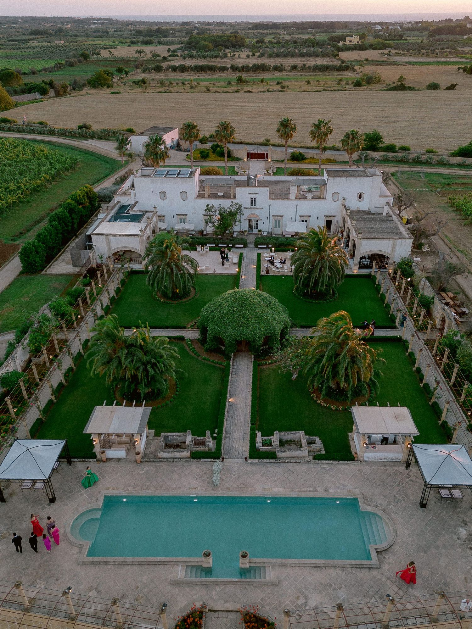 Aerial view of a luxurious Tenuta San Mose with white buildings, palm trees, manicured gardens, and a swimming pool in desert surroundings.