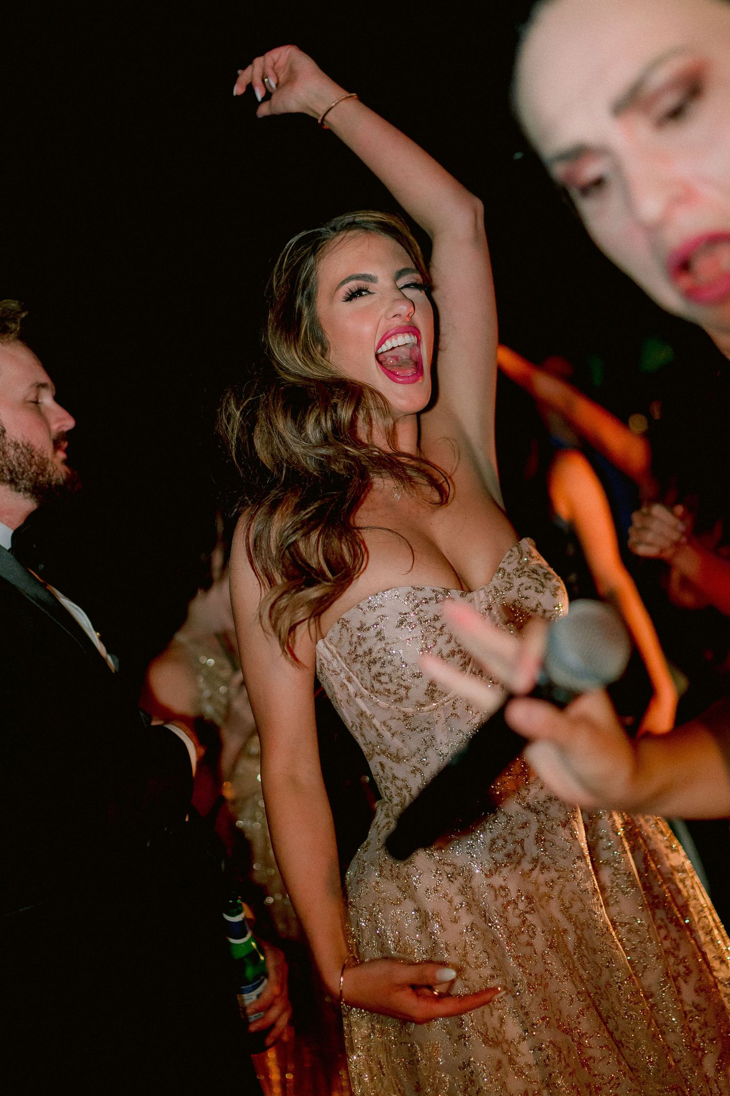Woman in sparkly champagne gown dances joyfully with arm raised at a festive nighttime celebration.