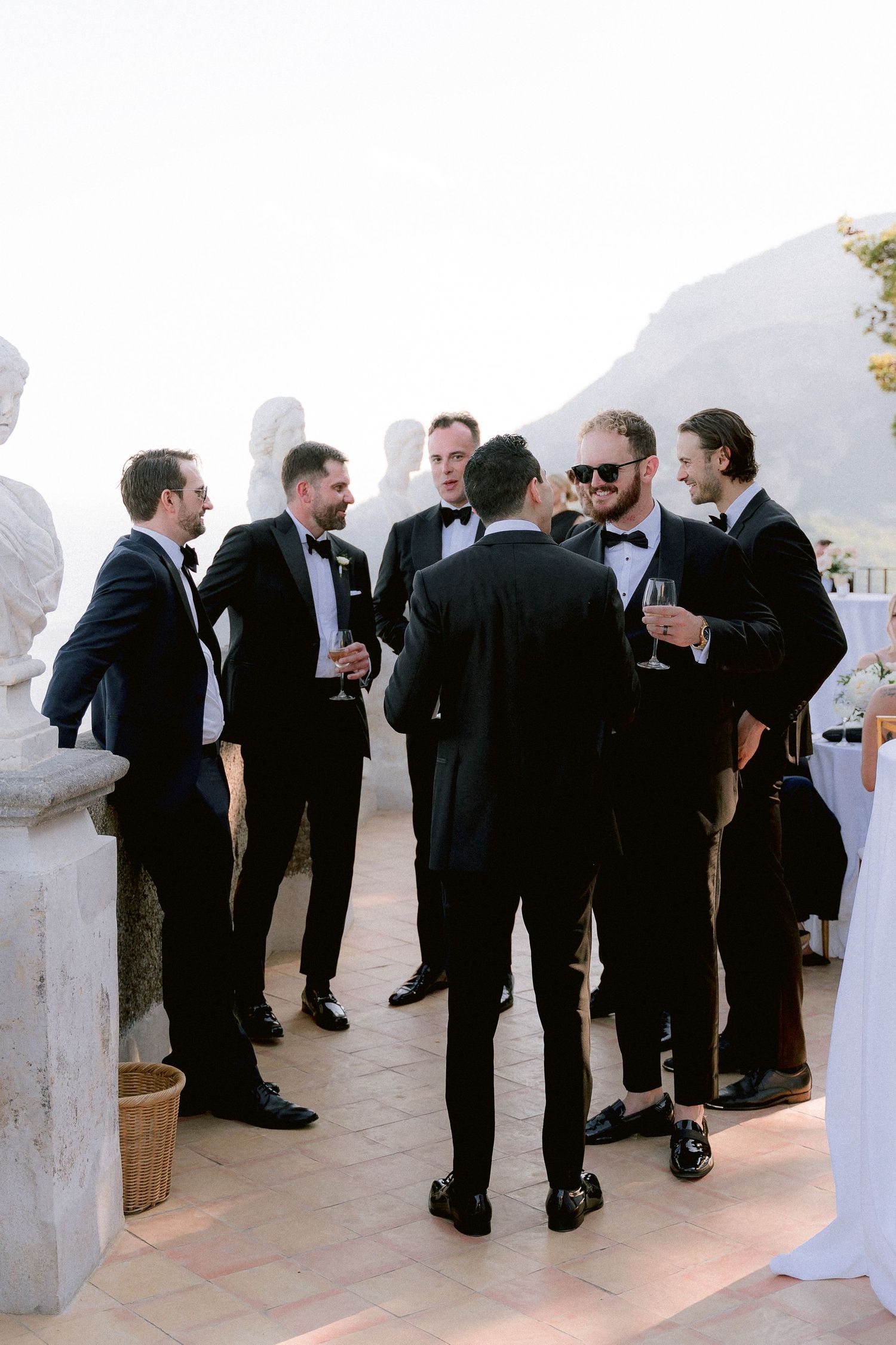 A group of groomsmen in formal black tuxedos and suits gather on a terrace for pre-wedding celebration drinks.