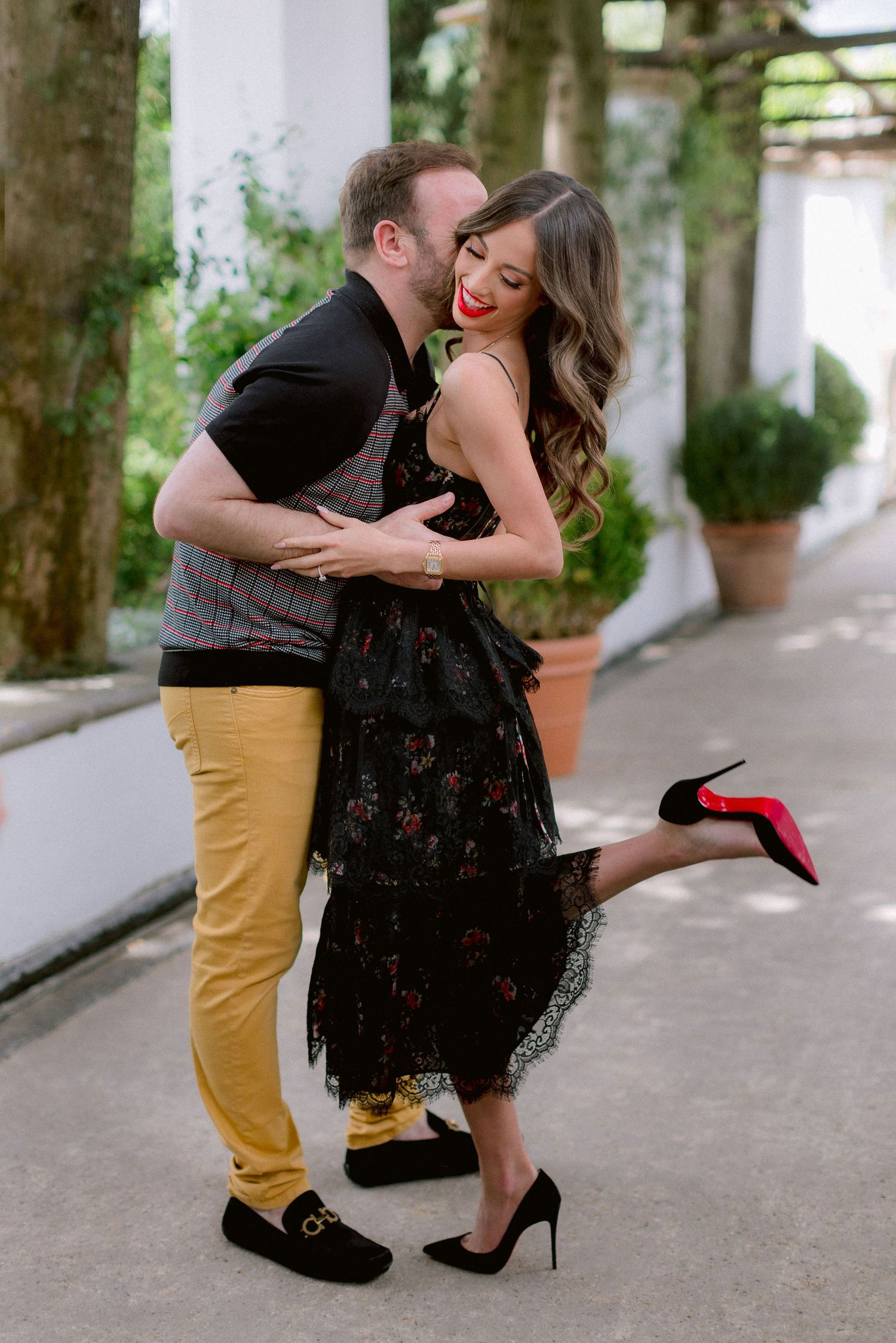 A romantic moment shared between two people in a garden setting, with her wearing a black lace dress and red heels.