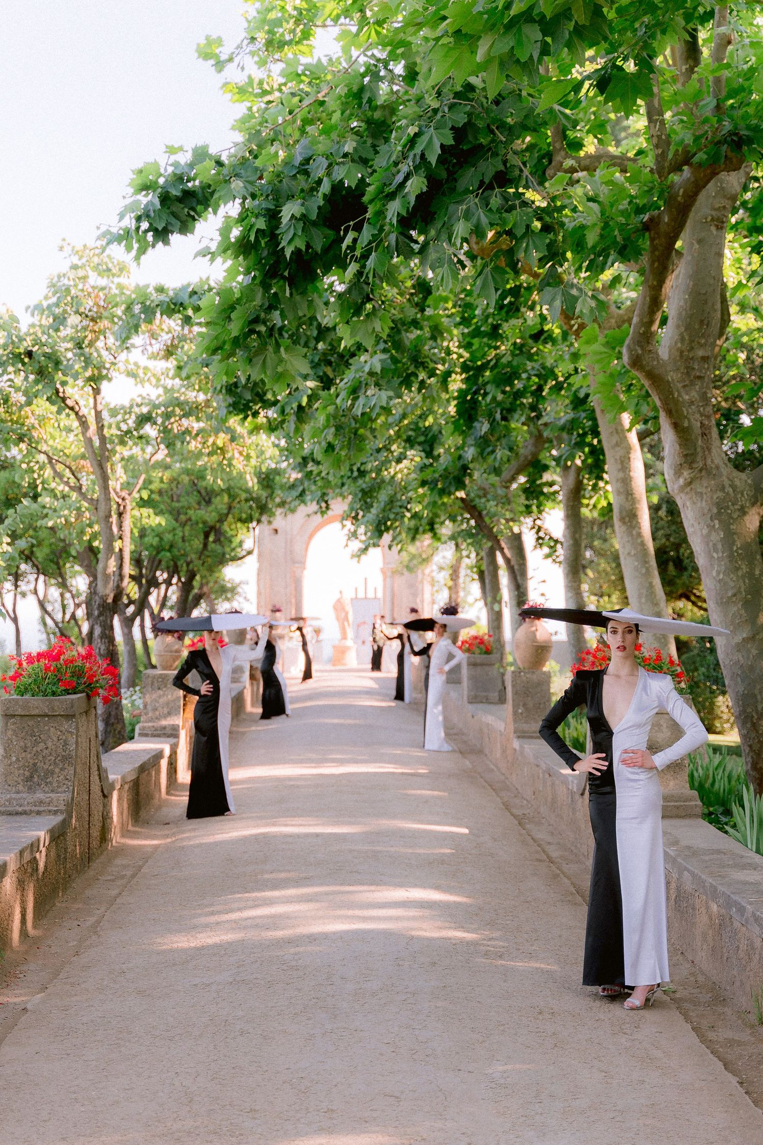 Elegant women in black and white dresses pose along a tree-lined pathway leading to terrazza dell'infinito in a Mediterranean garden.