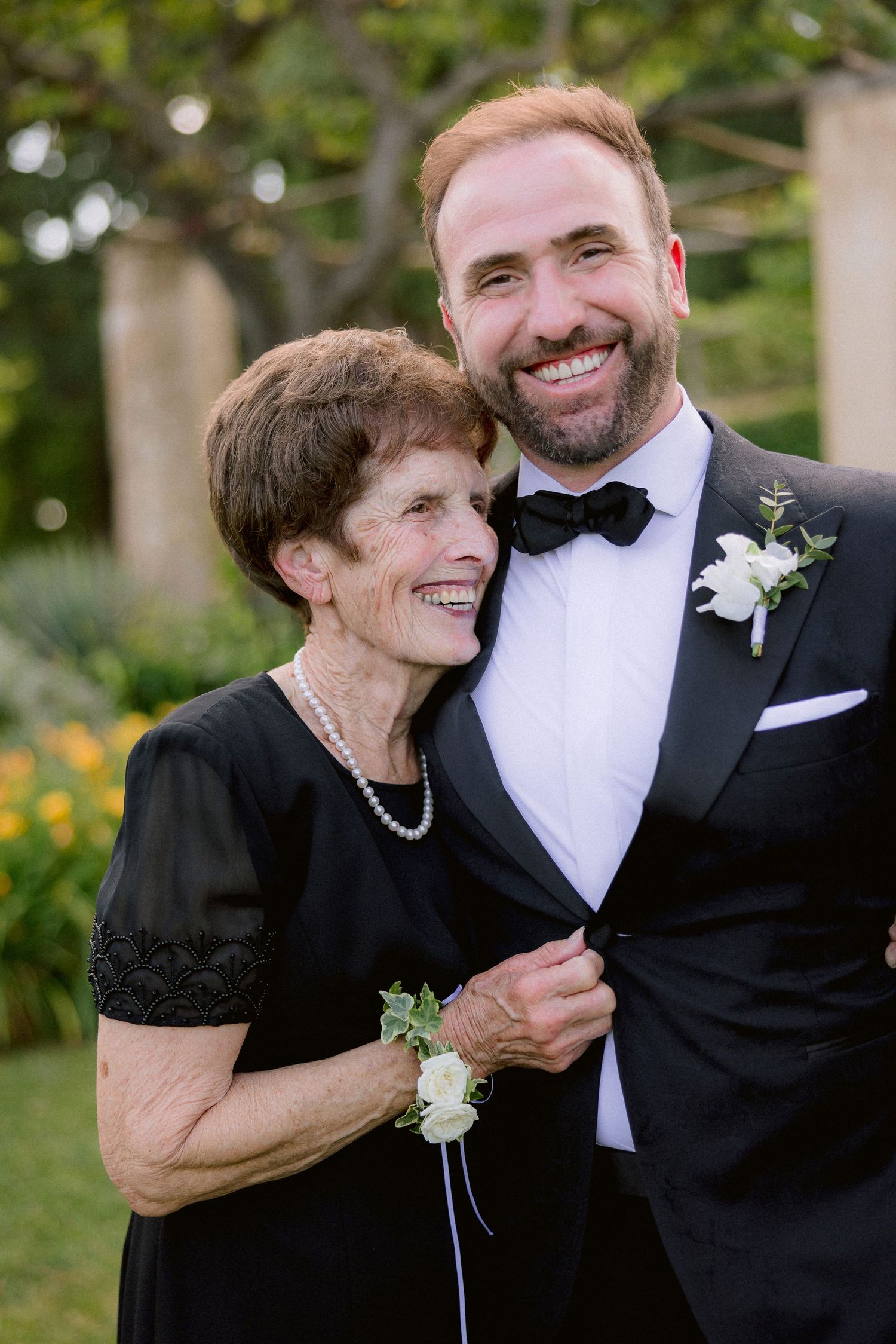 Two people in formal attire at a wedding, wearing black with boutonnieres and smiling outdoors among greenery.