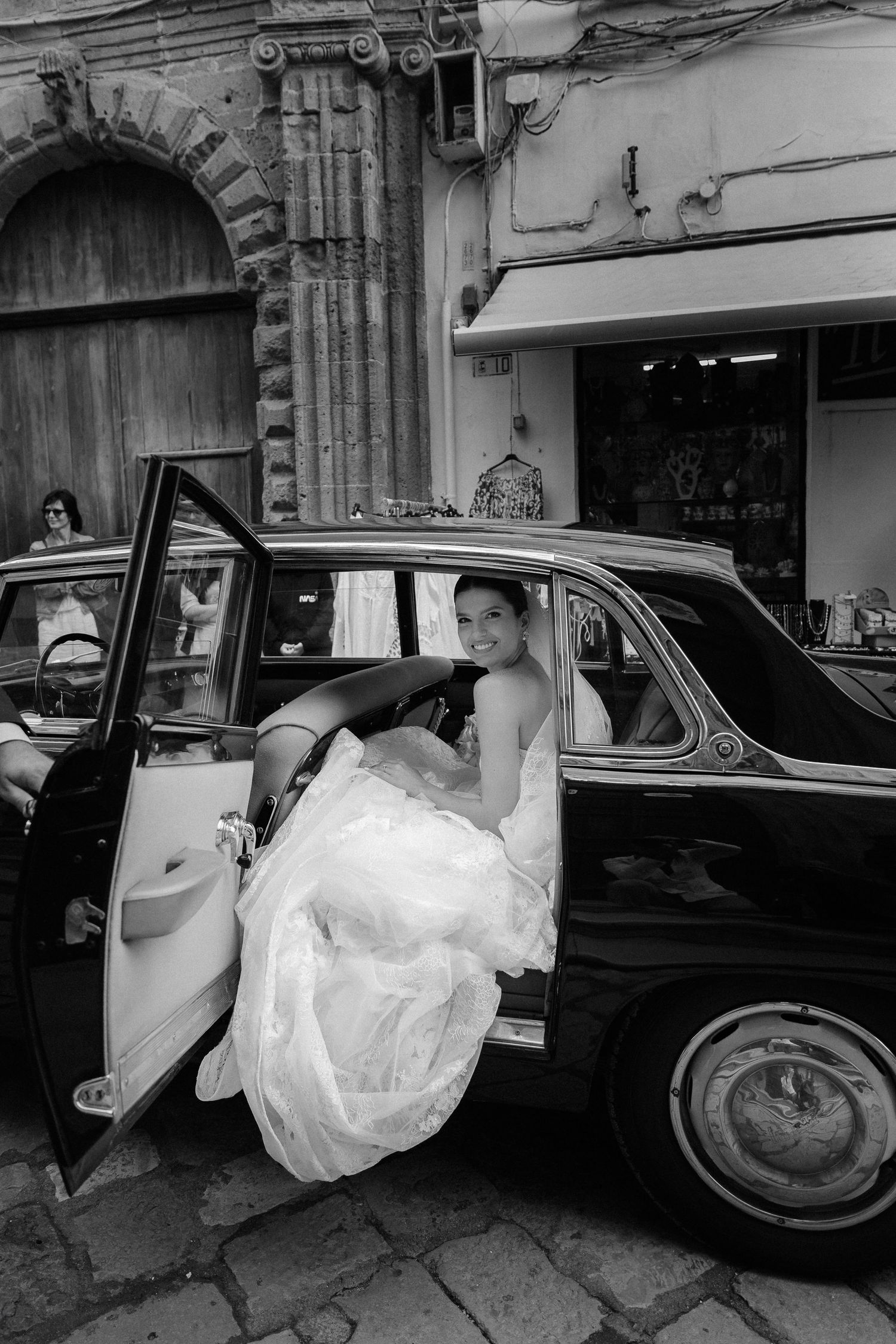 A vintage black car with open door showcasing an elegant white wedding dress against a historic stone building facade in Gallipoli.