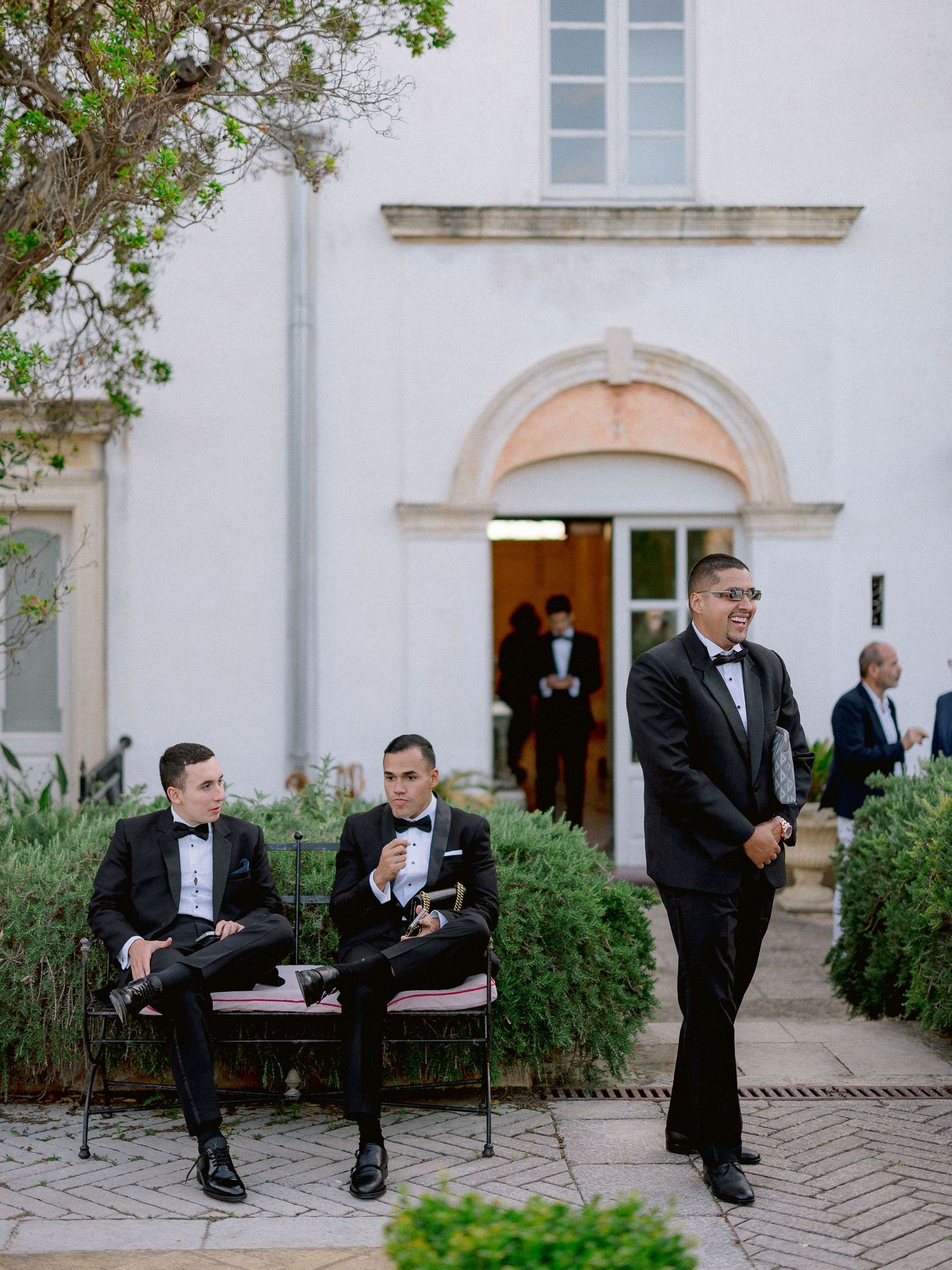 Members of the wedding party relax on a bench and stand near an elegant white church entrance with arched doorway.