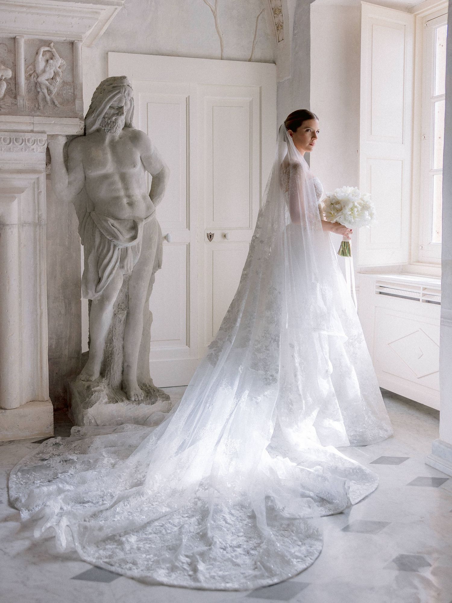 Elegant bridal scene with flowing lace wedding gown and cathedral veil next to classical marble statue in white room.