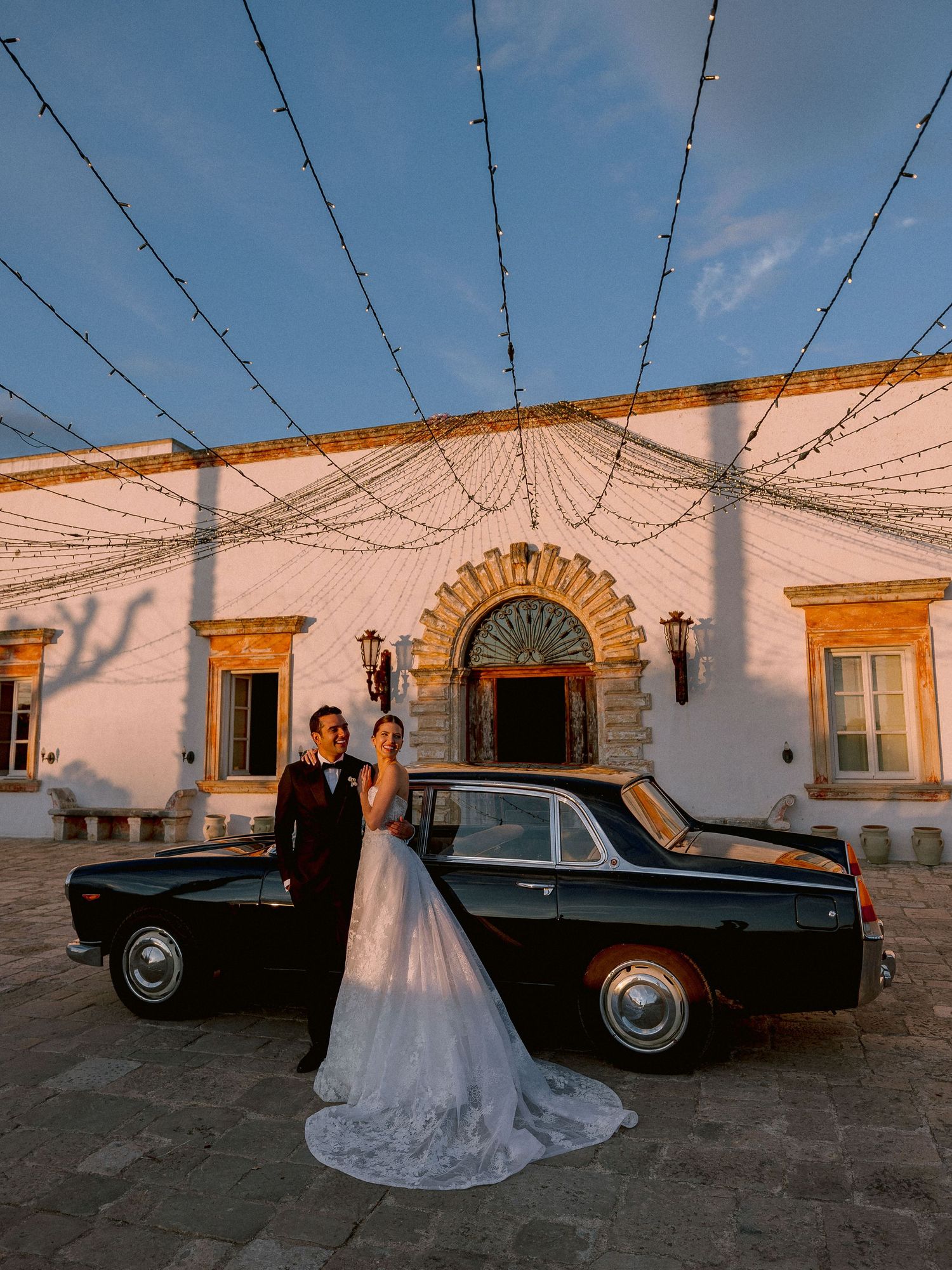 A vintage black car parked in front of Tenuta San Mose in Puglia with string lights and an ornate arched entrance at sunset.