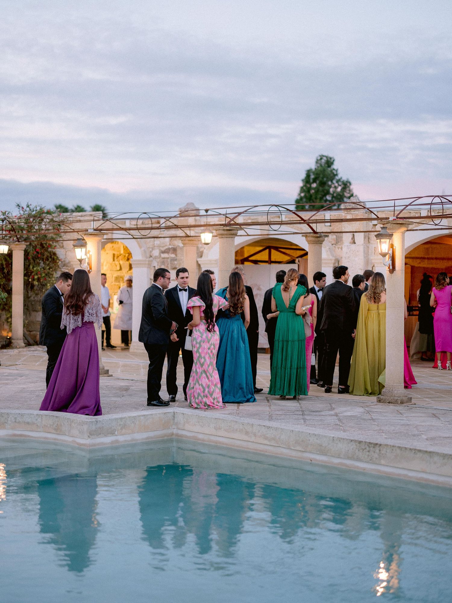 A group of elegantly dressed guests gather around a pool at a luxury venue during sunset for a formal event.