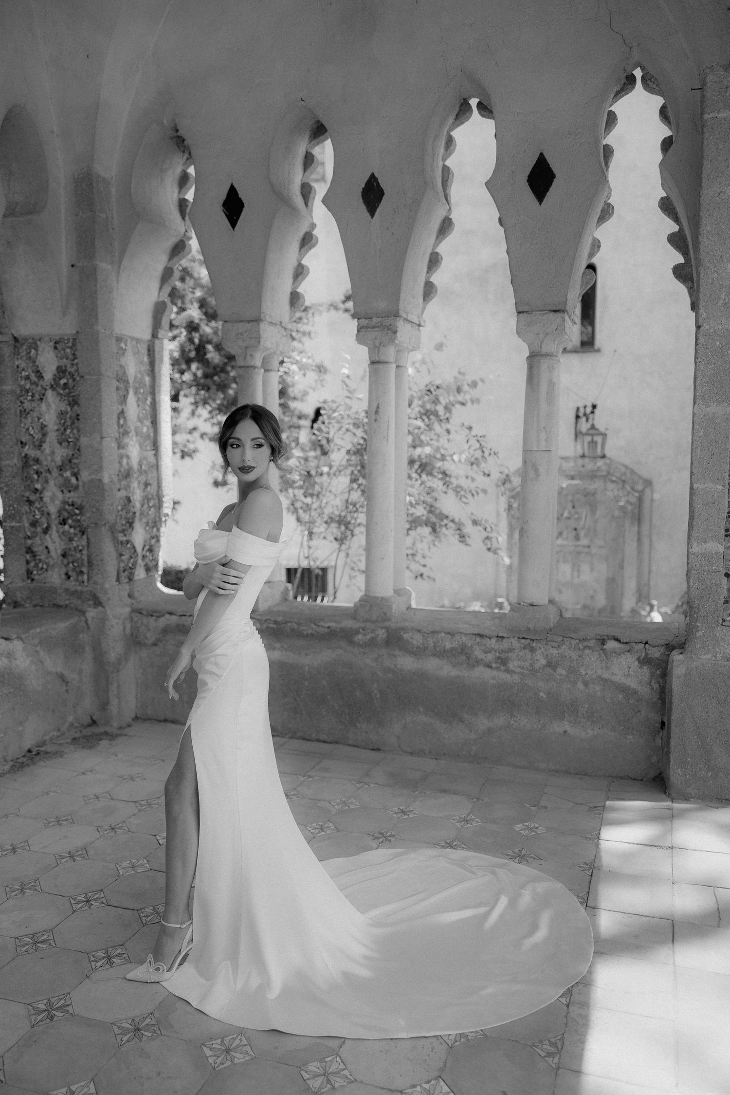 A bride in an elegant off-shoulder white gown poses in a historic stone archway with gothic architectural details.