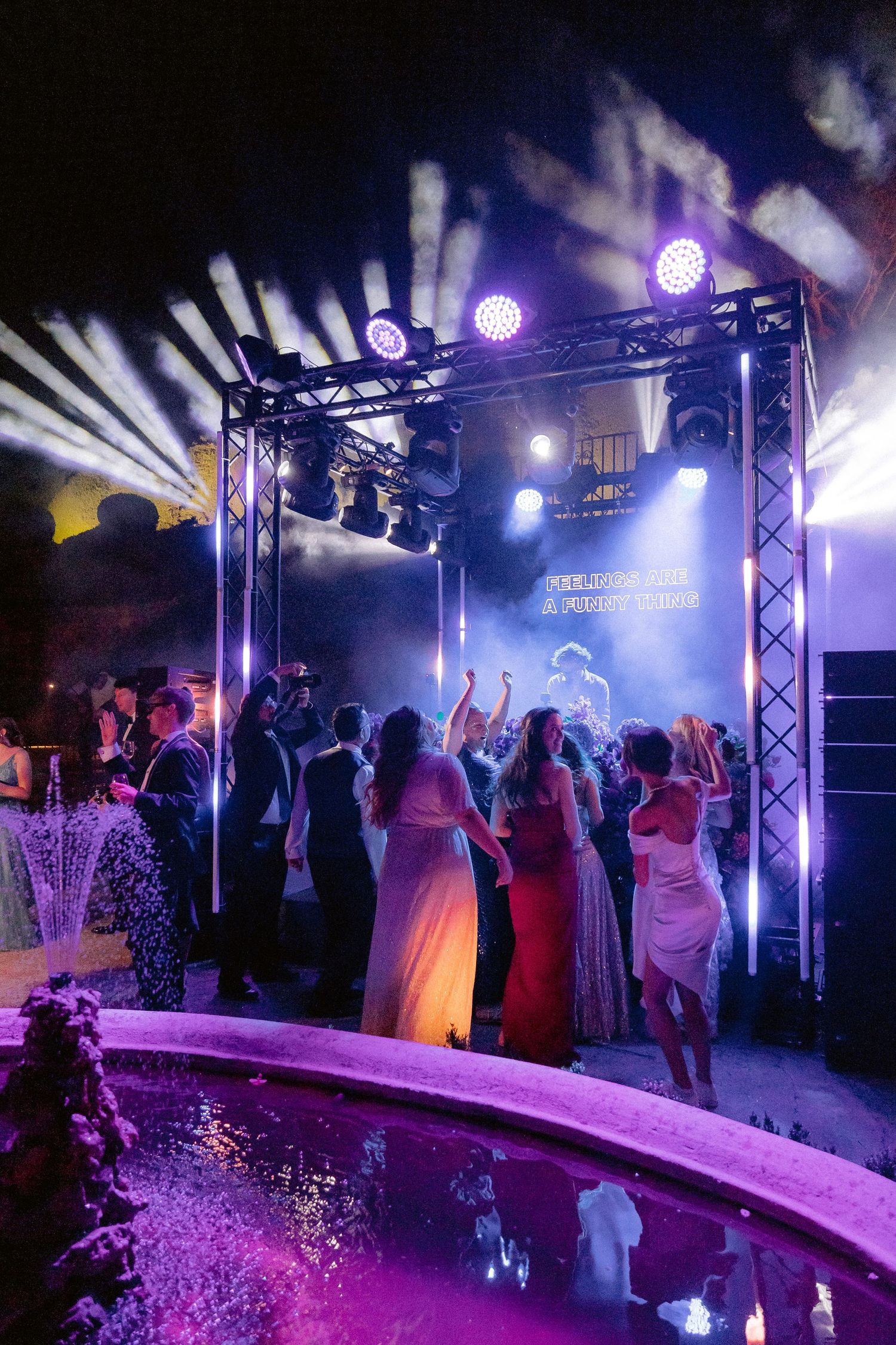 Party guests dance at an outdoor wedding reception with purple lighting and stage truss setup near a curved pool.