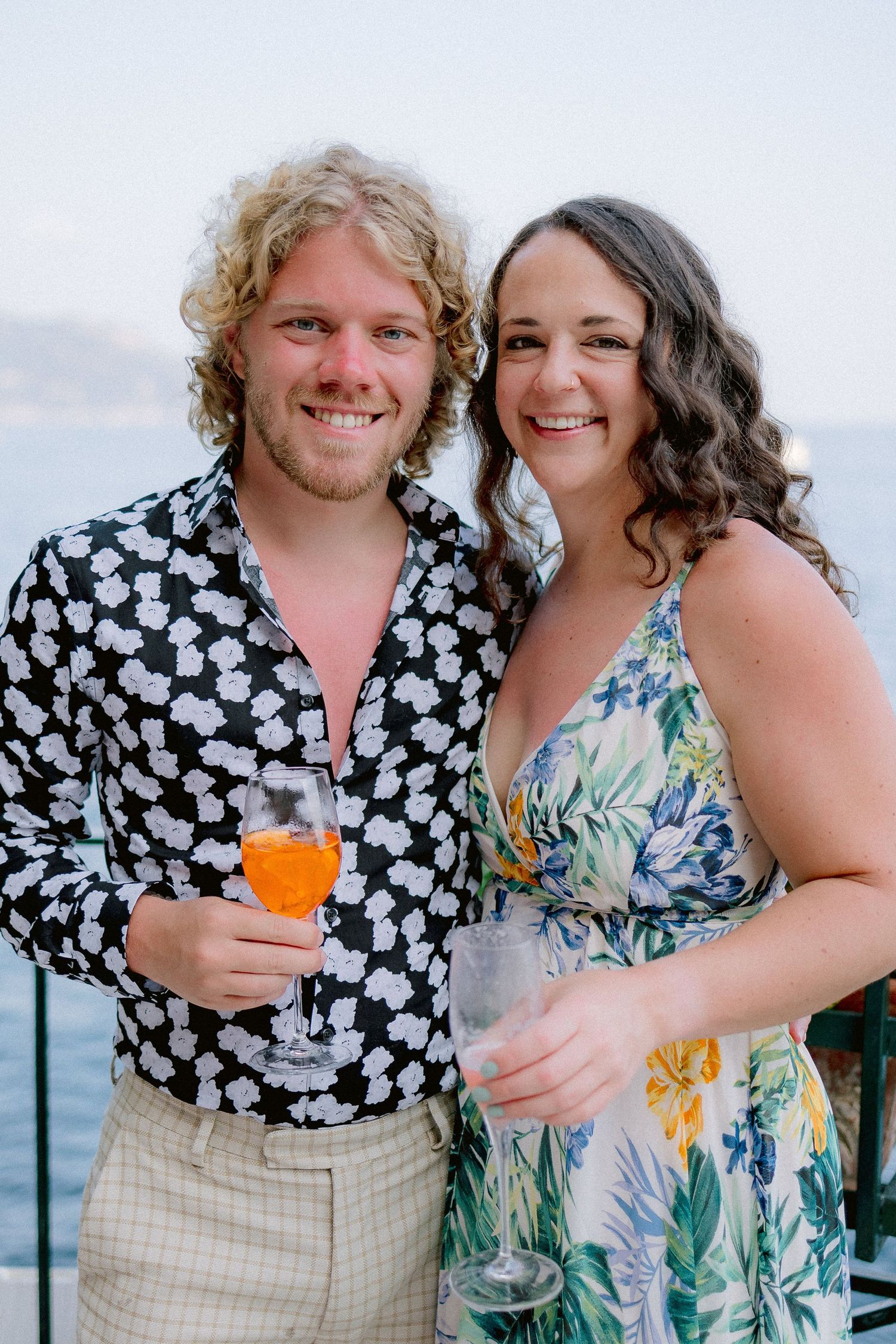 A couple in tropical attire pose together holding cocktails against a seaside backdrop.