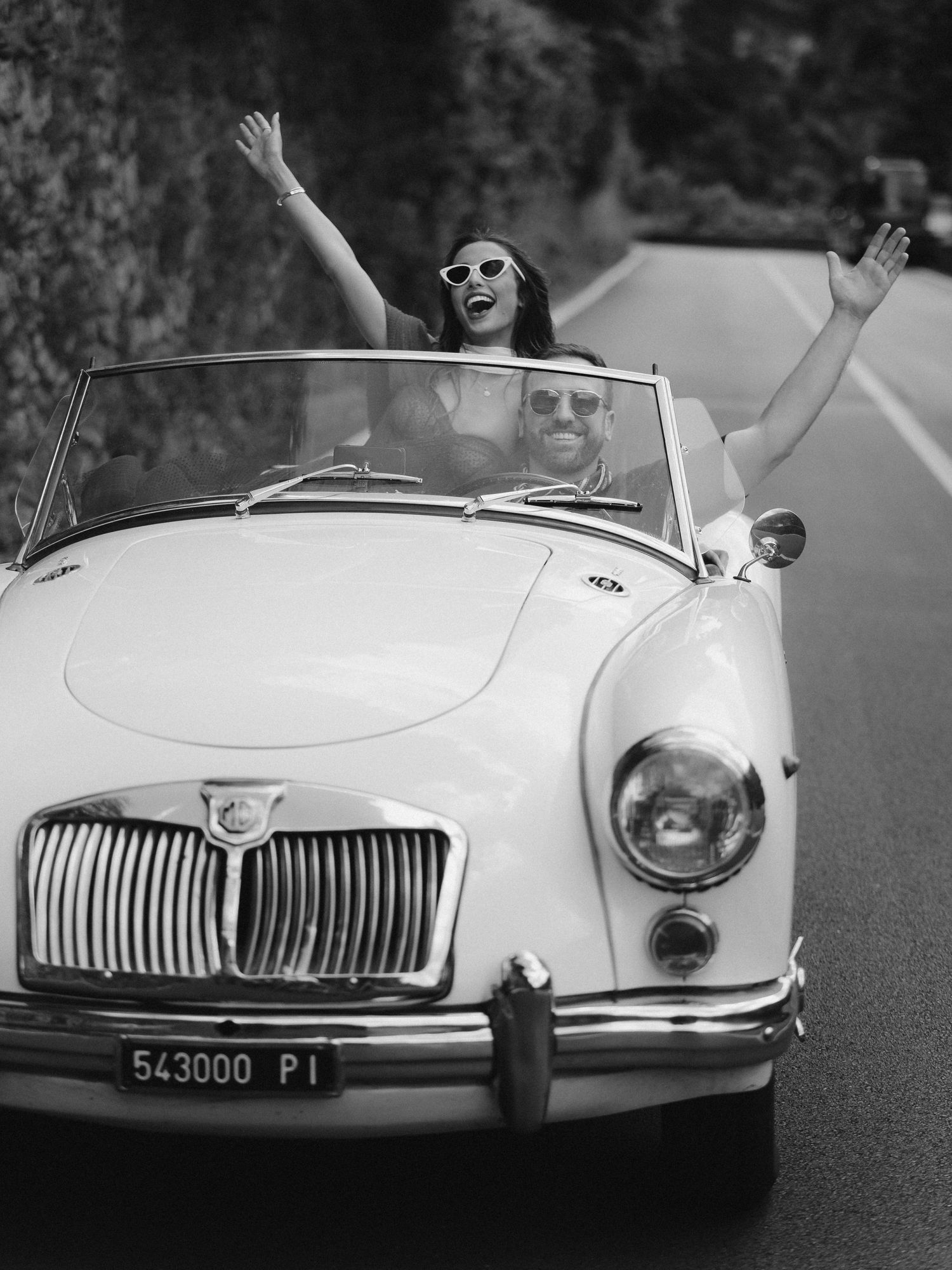Black and white photo of two people enjoying a carefree ride in a classic white MG convertible on a winding road.