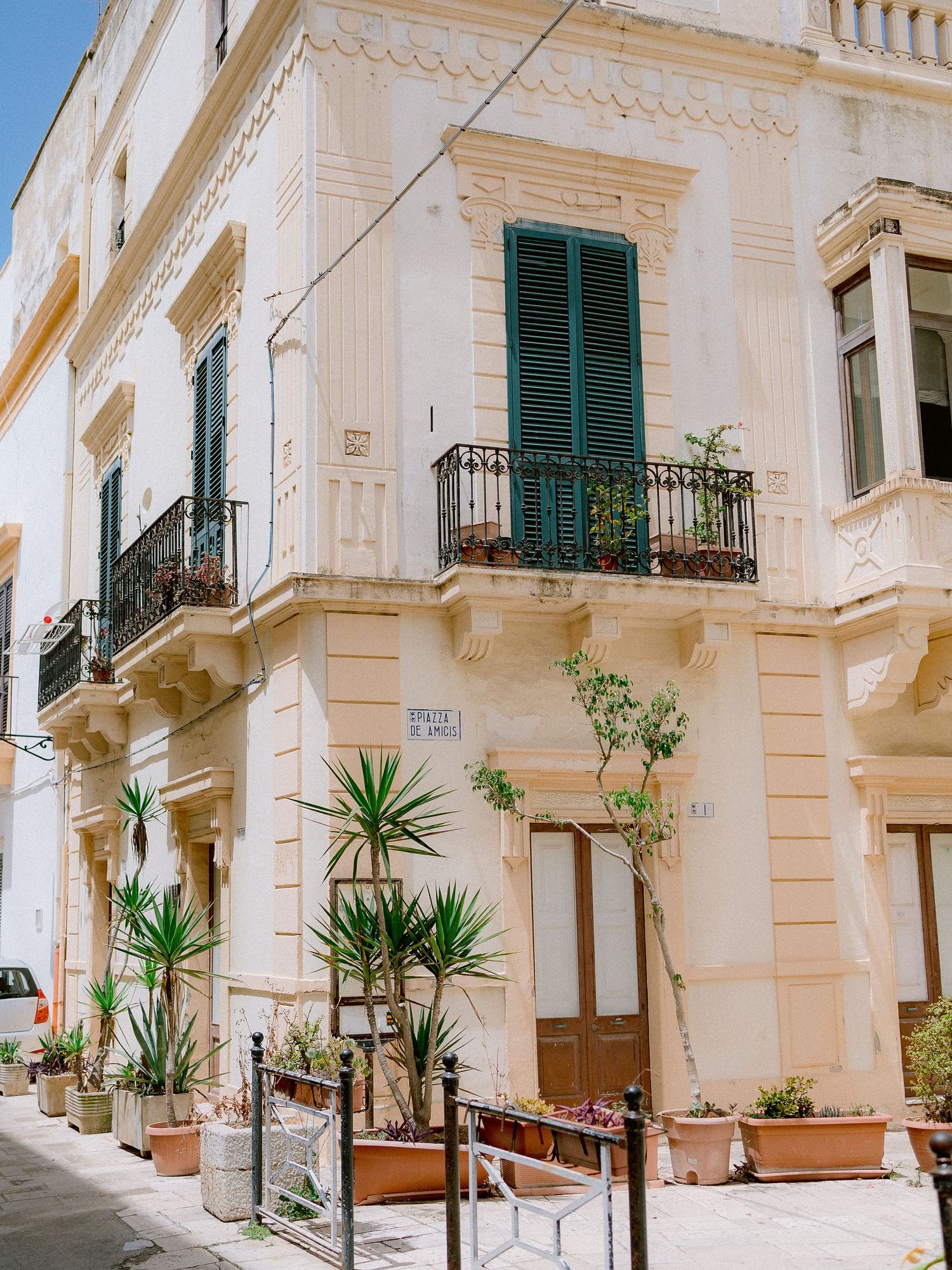 Mediterranean style corner building in Gallipoli with white walls, green shutters, ornate balconies and potted palm plants along sidewalk.