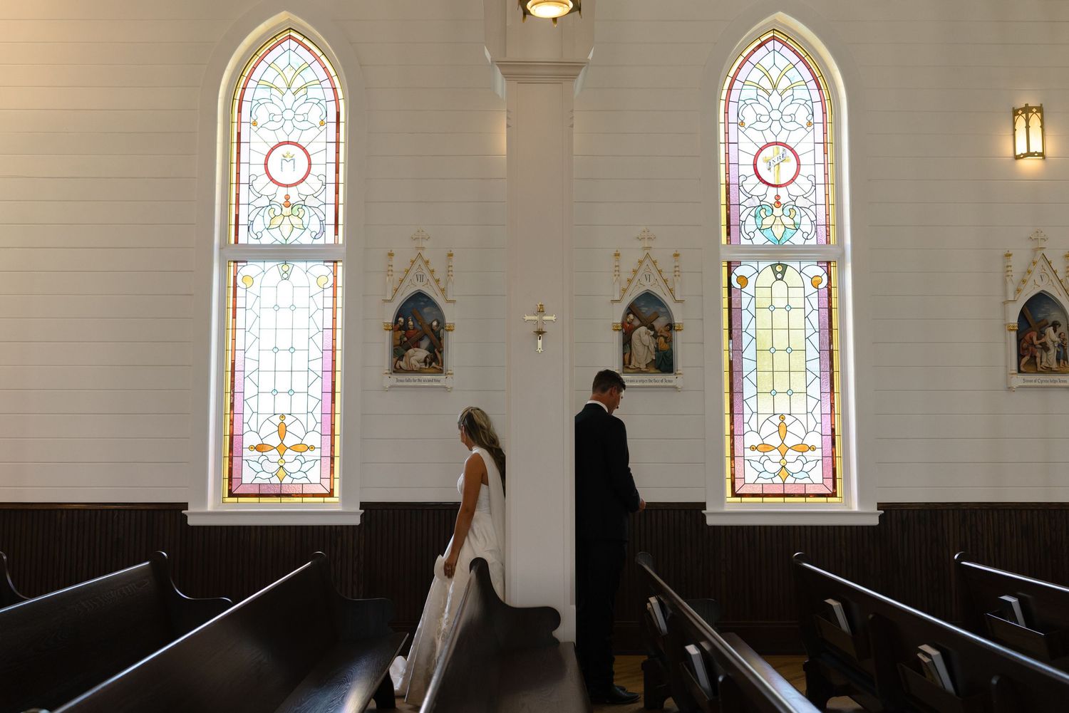 Two people stand in a church aisle between wooden pews with stained glass windows and religious statues on the walls.