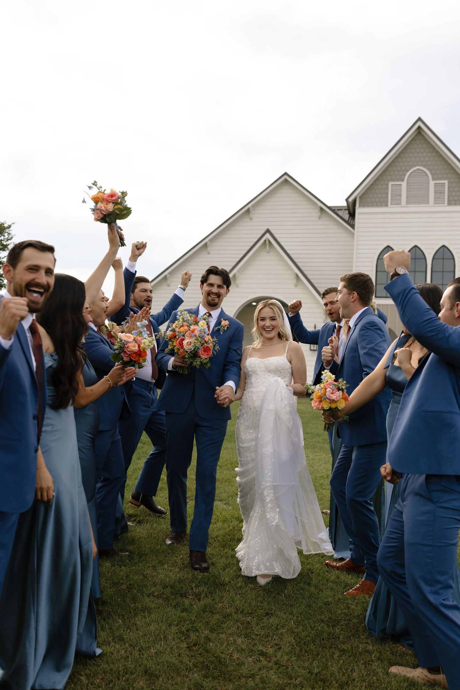 A joyful wedding celebration on a grassy lawn outside a white church with the wedding party wearing navy blue attire.