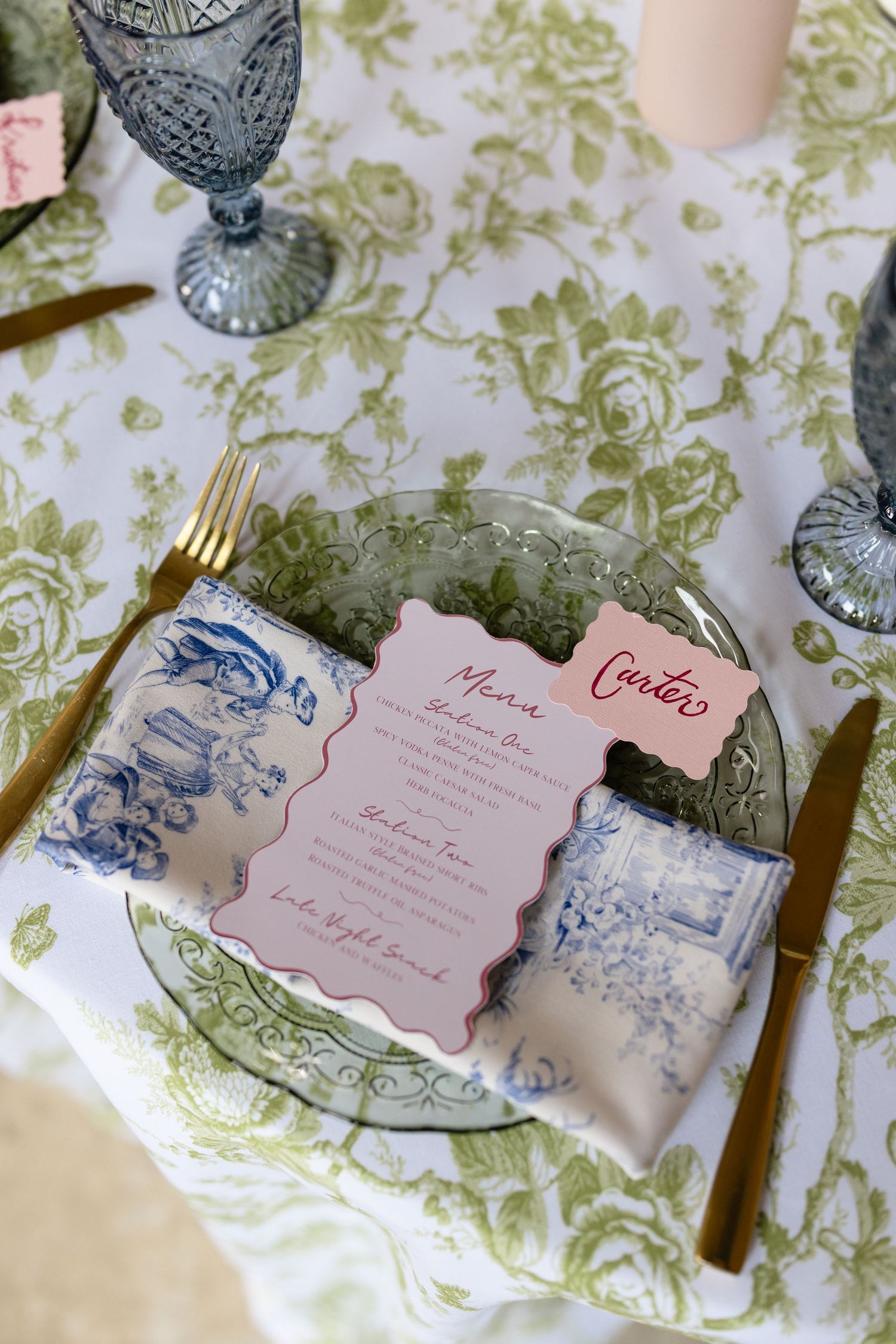 Elegant place setting with pink menu card on green floral tablecloth with blue vintage glassware and china patterns.