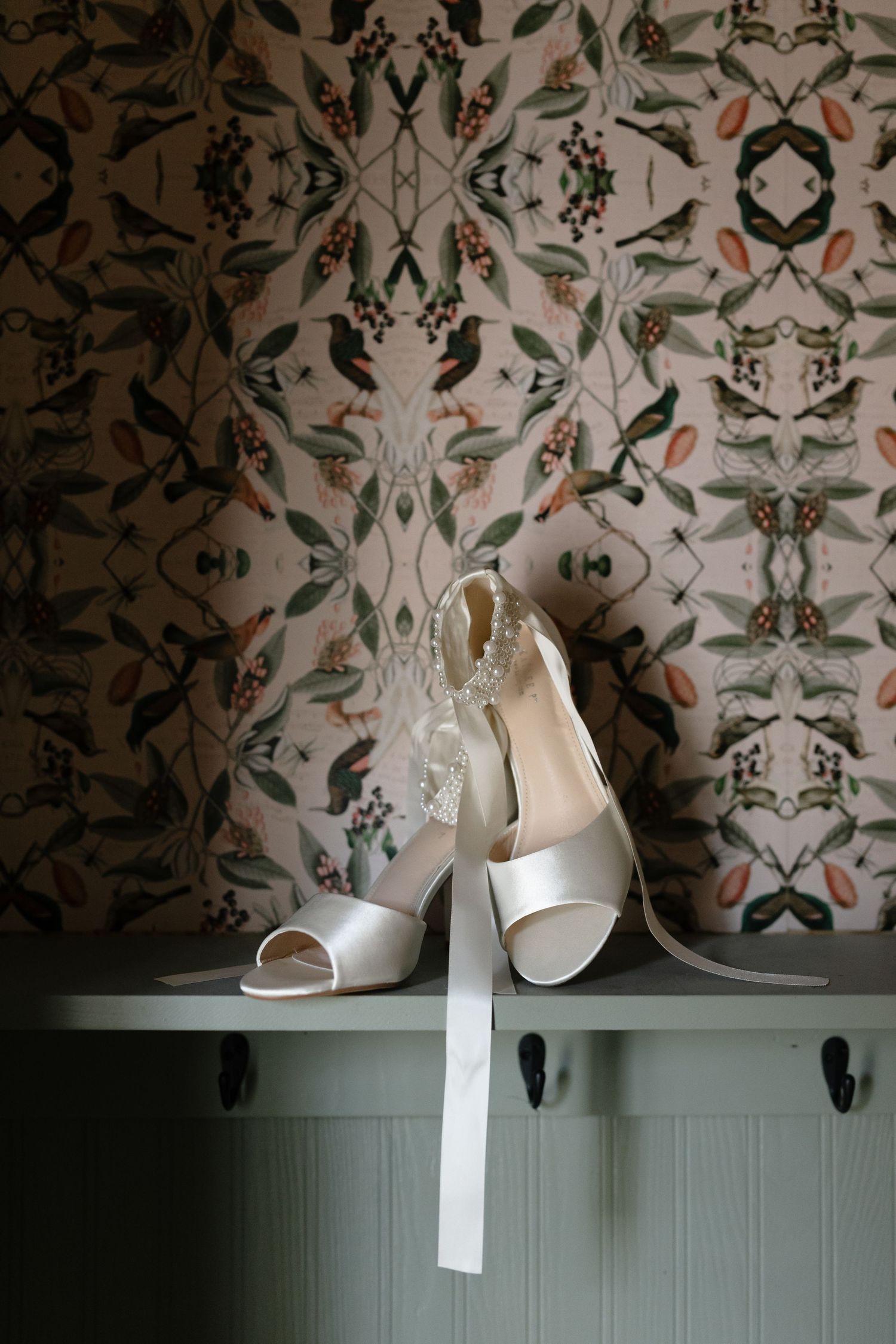 White wedding shoes hanging on black hooks against a floral wallpaper background in a boutique setting.