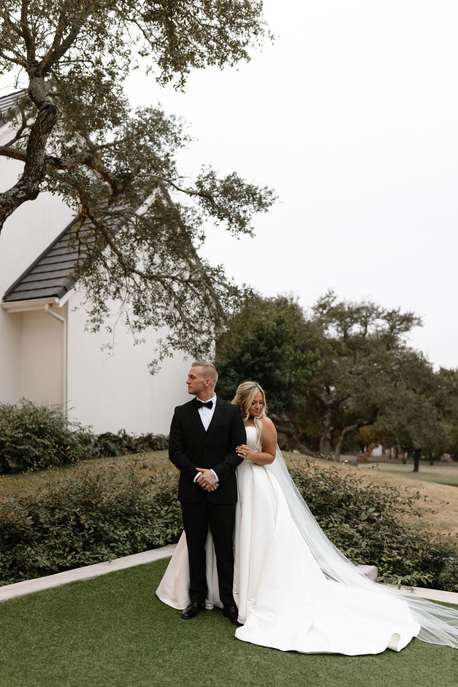 A couple in formal wedding attire stands together on a manicured lawn next to a white church building with oak trees behind them.