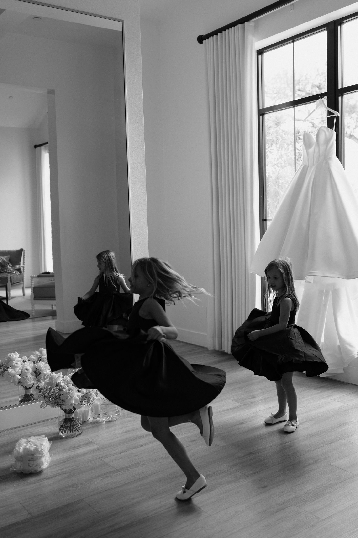 Black-clad dancers twirling in a bright room with hardwood floors as a white wedding dress hangs by the window.