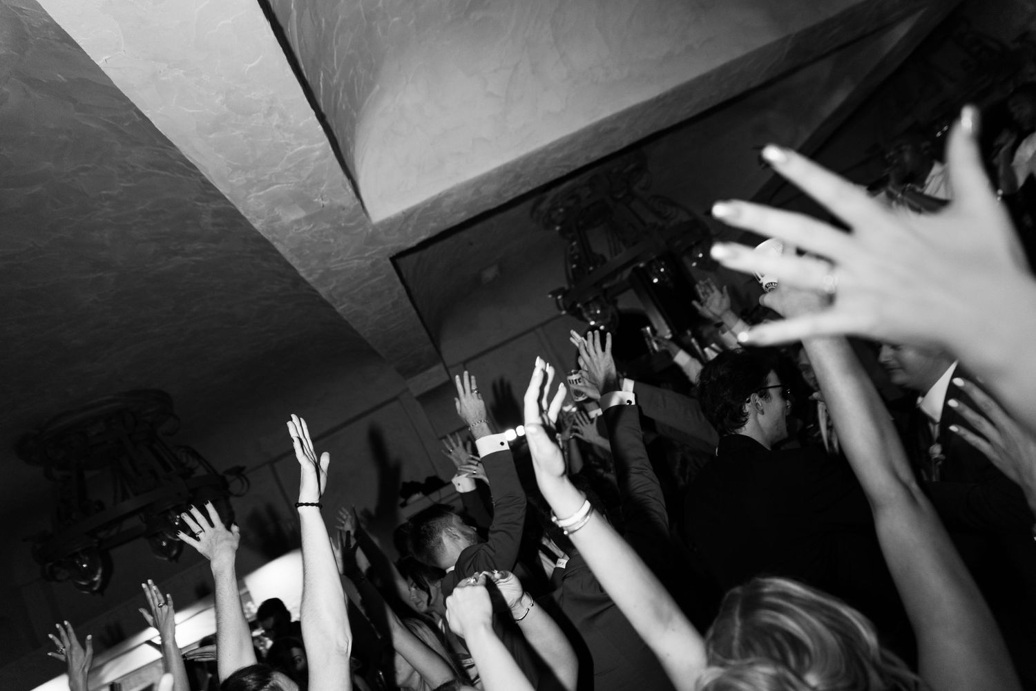 Black and white photo of raised hands at a concert venue with crowd reaching upward in enthusiastic celebration.