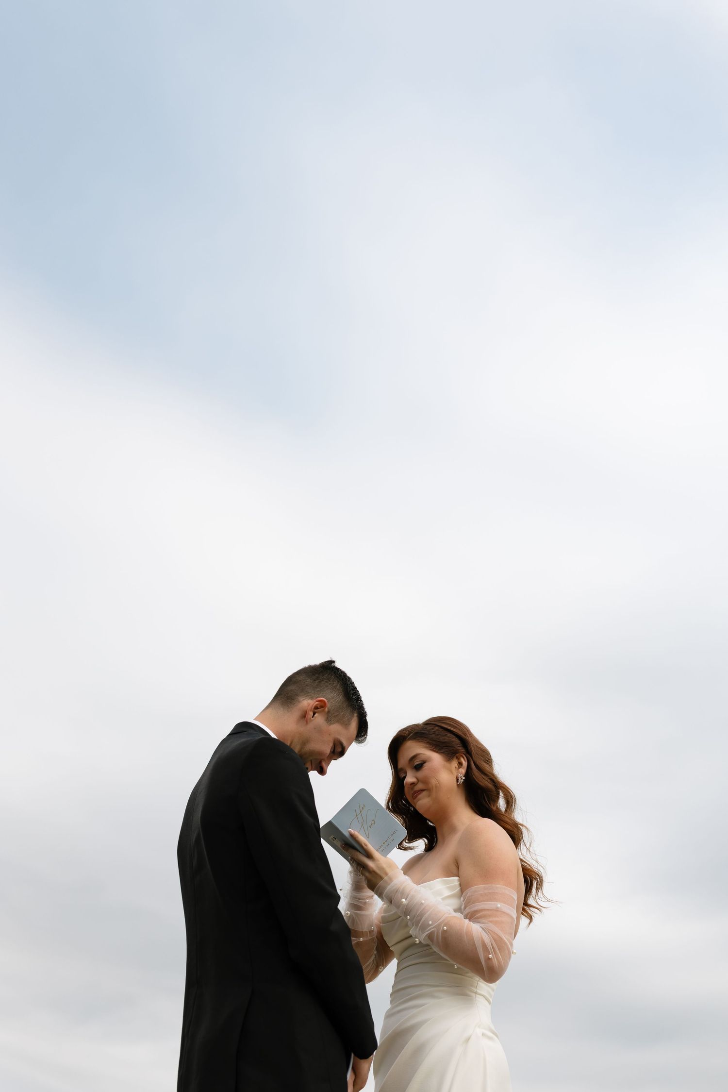 A couple in formal wedding attire share an intimate moment together outdoors against a cloudy sky backdrop.
