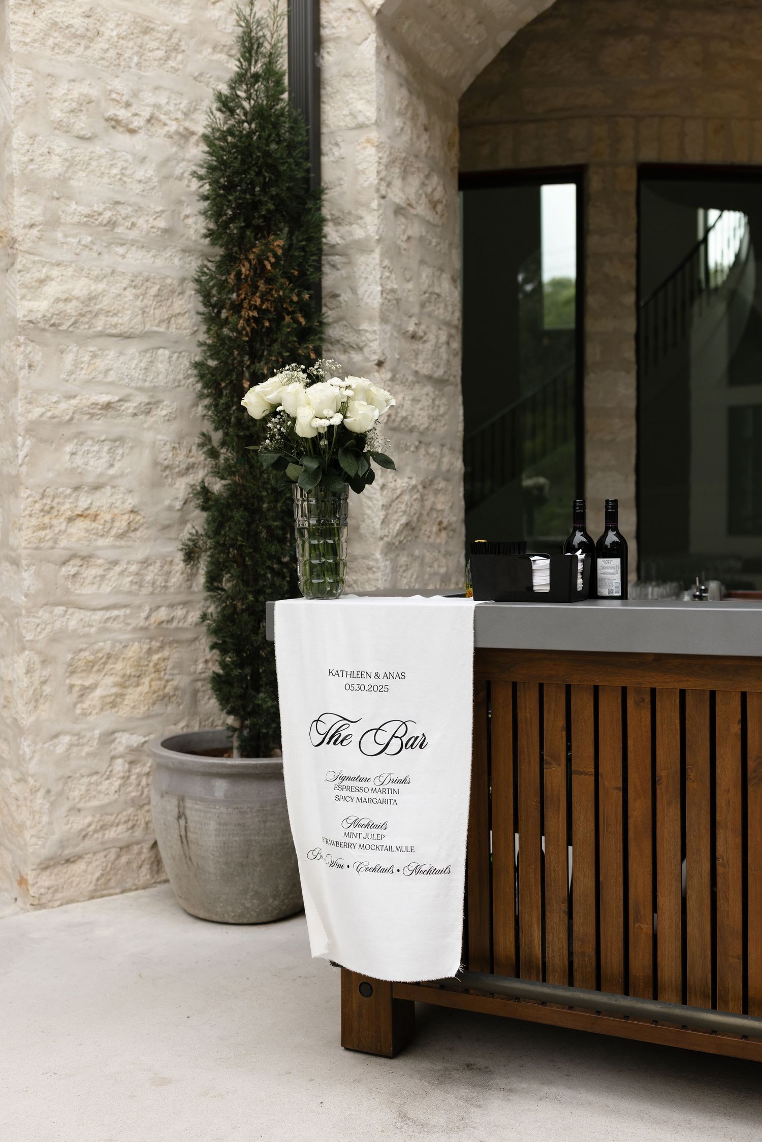 An elegant bar setup with white roses in a vase, wine bottles, and a white sign reading 'The Bar' against a stone wall.