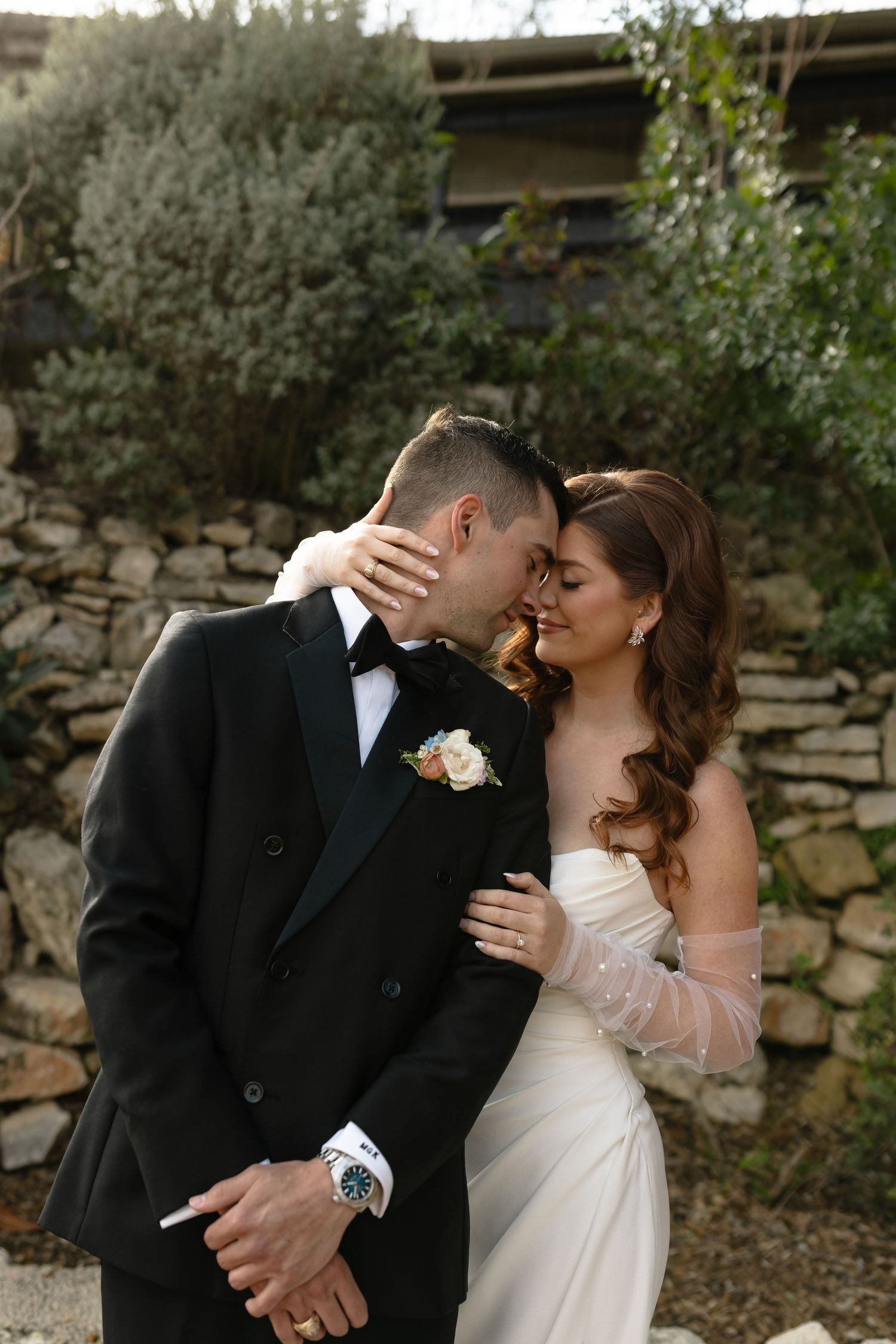 A romantic wedding couple share an intimate moment against a rustic stone wall backdrop at sunset.
