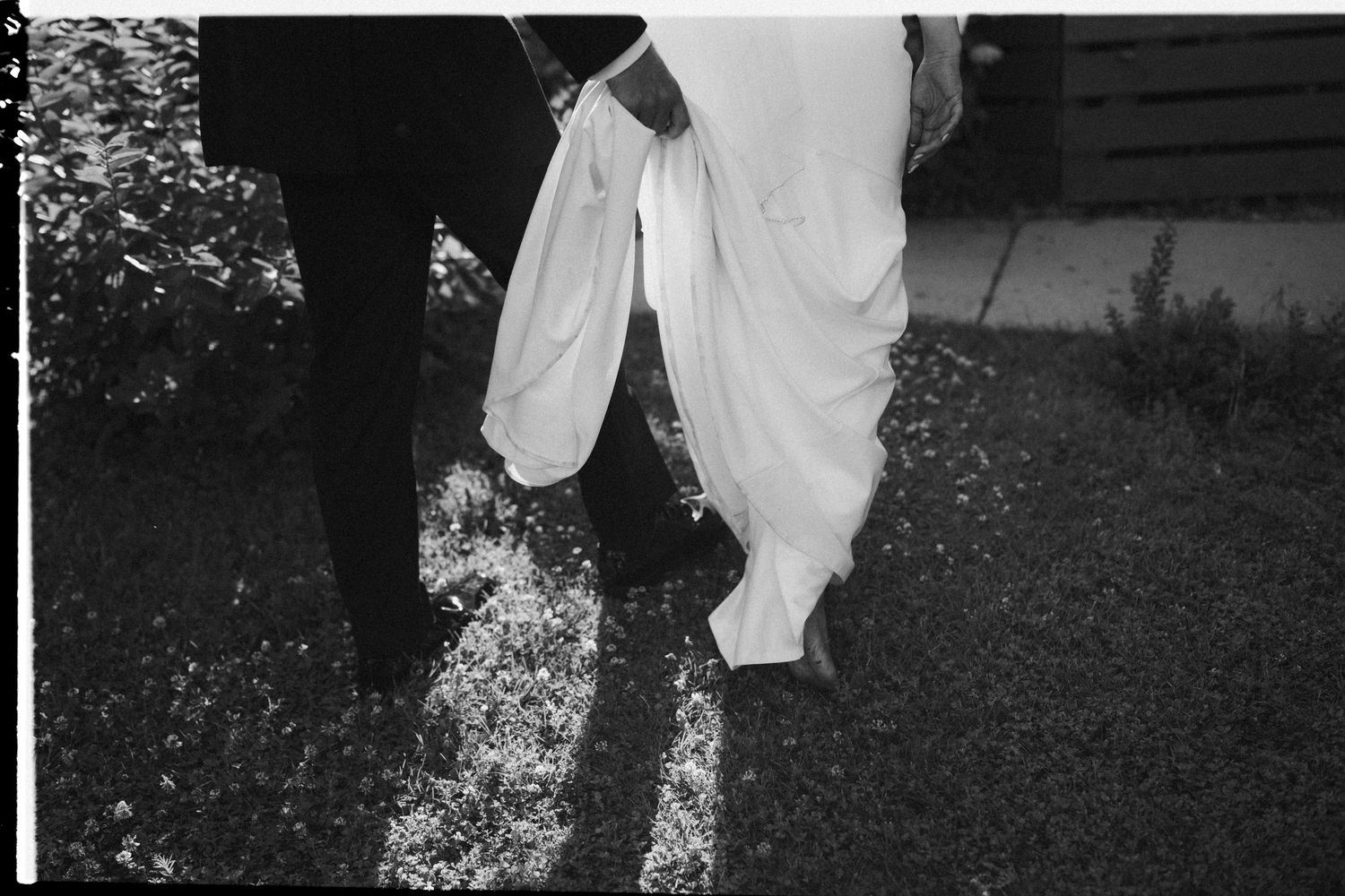Person in a garden sniffs a white flower while wearing a white dress in a black and white photograph.