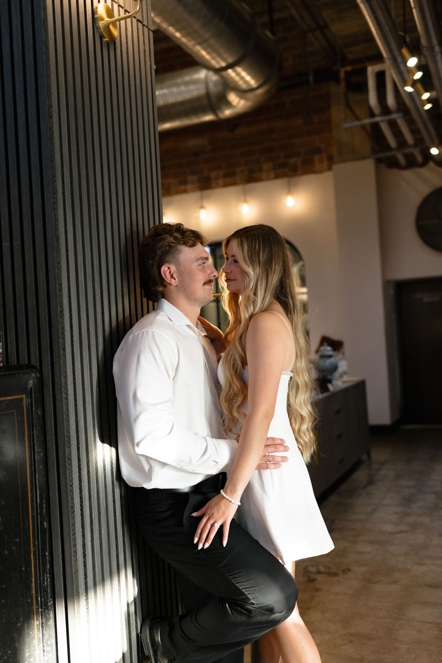Romantic couple in formal attire share an intimate moment against a dark wall in an industrial-style venue.