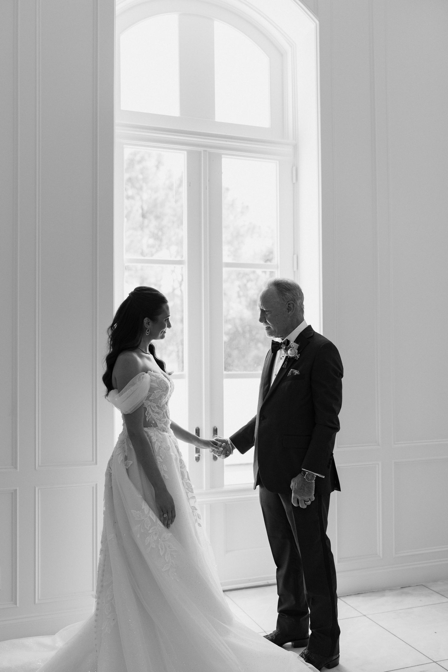 A tender black and white wedding moment between bride and her companion by a bright arched window in an elegant room.