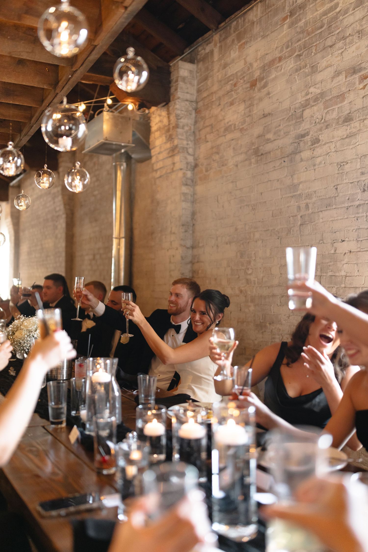 A lively group of bar patrons raise their drink glasses in a celebratory toast under rustic pendant lighting and exposed brick walls.