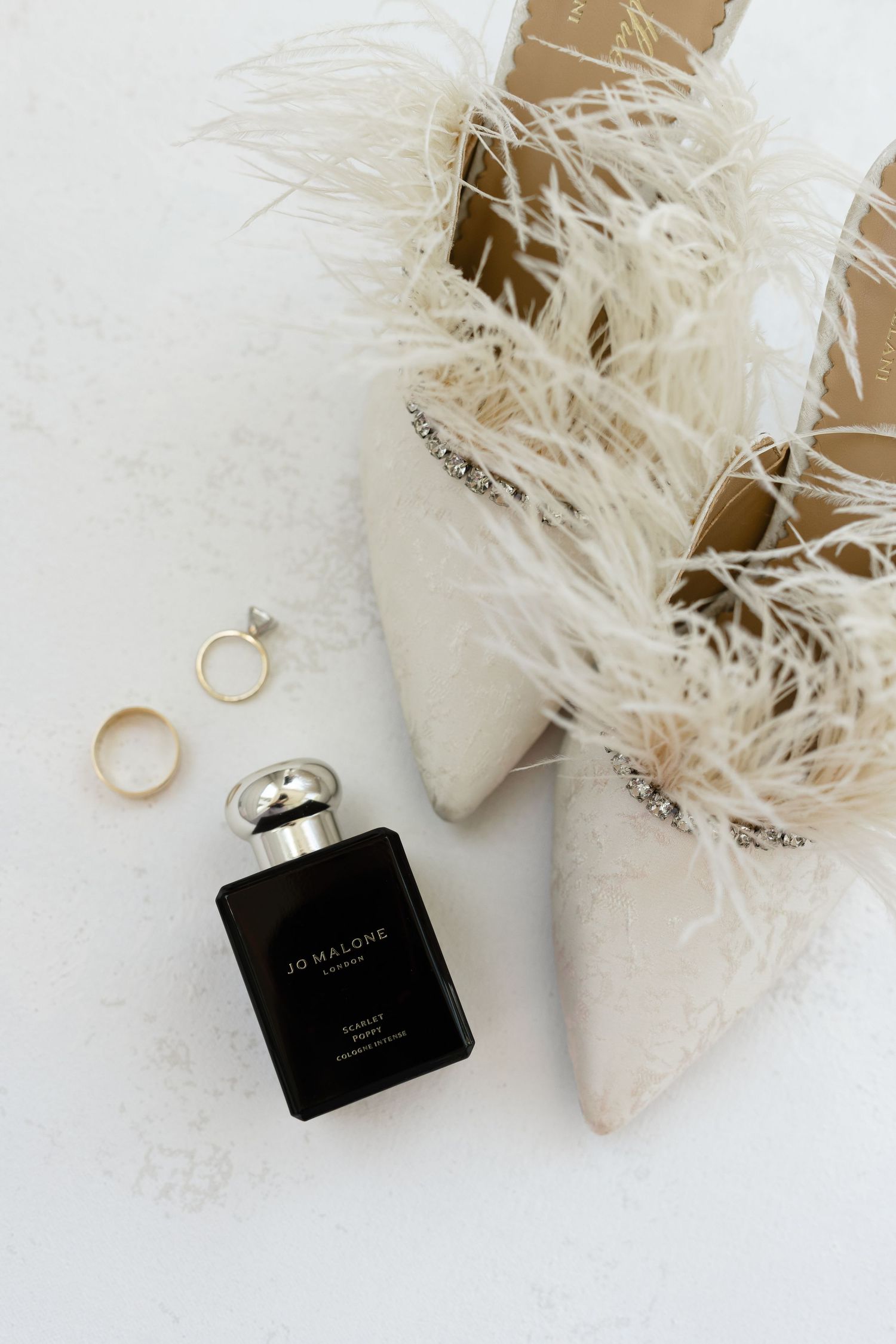 Wedding accessories including white feathered shoes, wedding rings and black Jo Malone perfume bottle arranged on white surface.