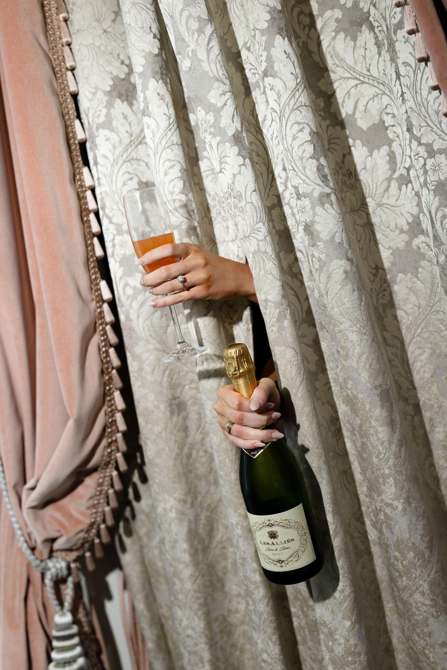 Hands reaching through elegant damask curtains holding a champagne bottle and flute against soft lighting.