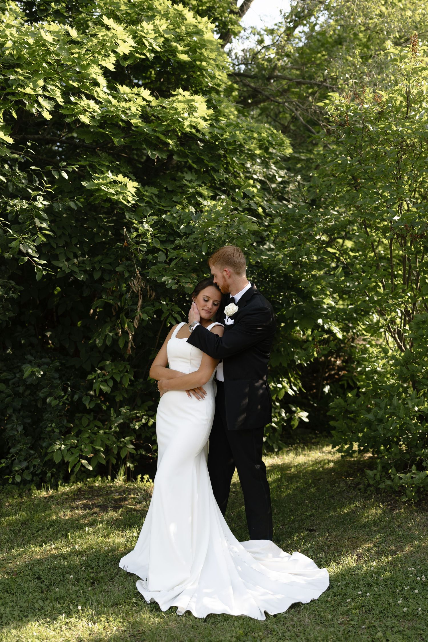 A couple shares a romantic moment in their wedding attire among green foliage on a sunny day.