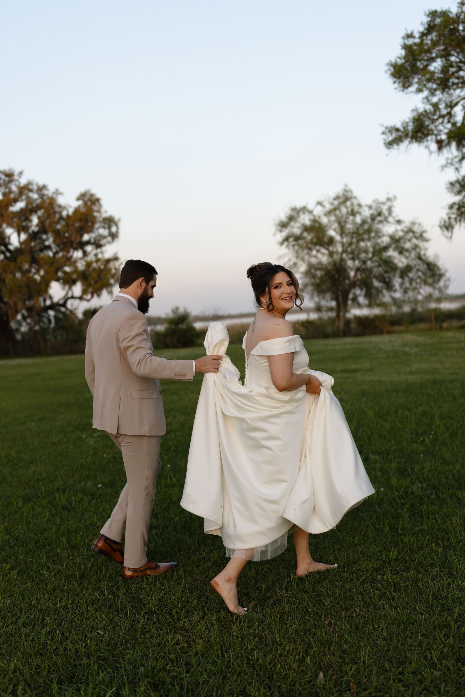 A couple shares an intimate moment dancing barefoot on grass at sunset in elegant wedding attire.