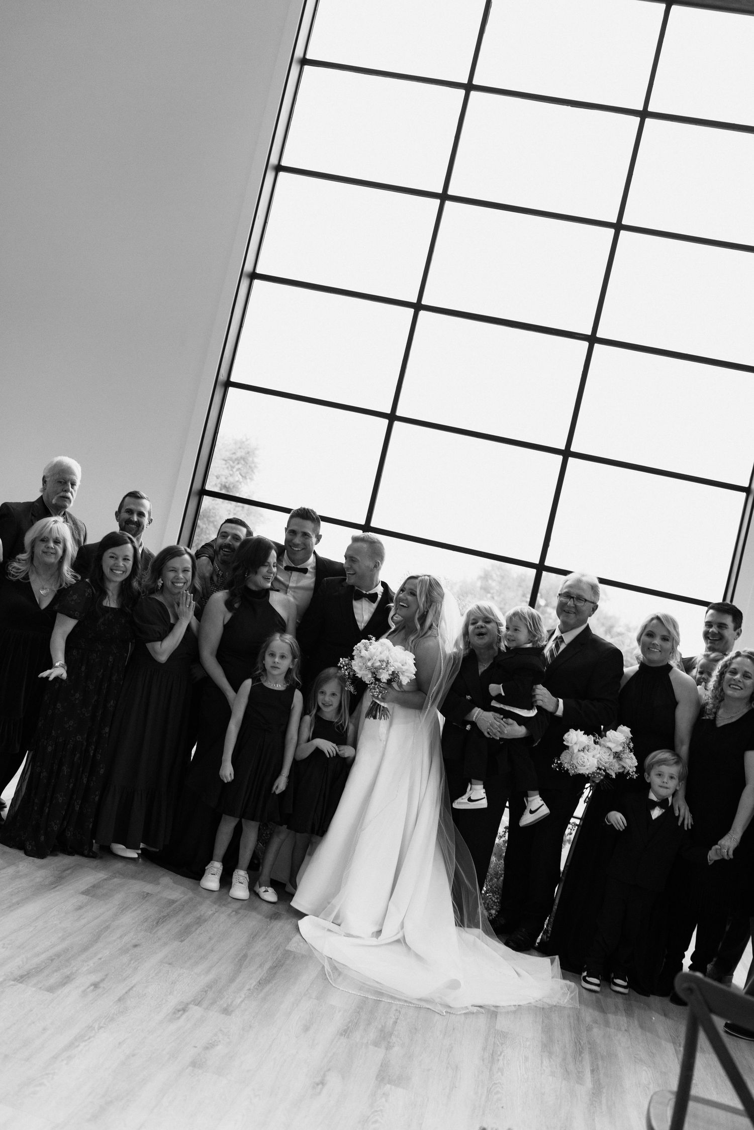 A wedding party poses together in black formal attire against a bright window-lit backdrop in a modern venue.