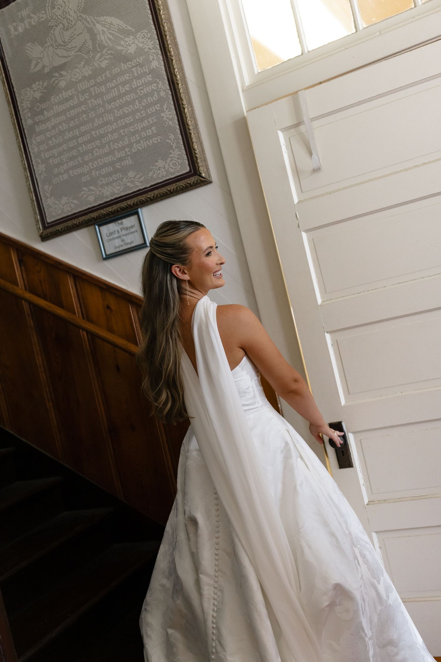 A person in a flowing white wedding dress stands by a wooden staircase and white door, turning to look back.