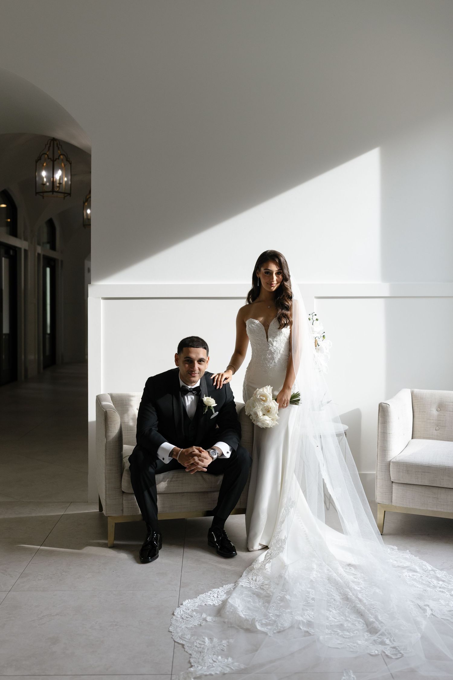 A bride in white lace gown and groom in classic tuxedo pose together against a white wall with dramatic lighting.